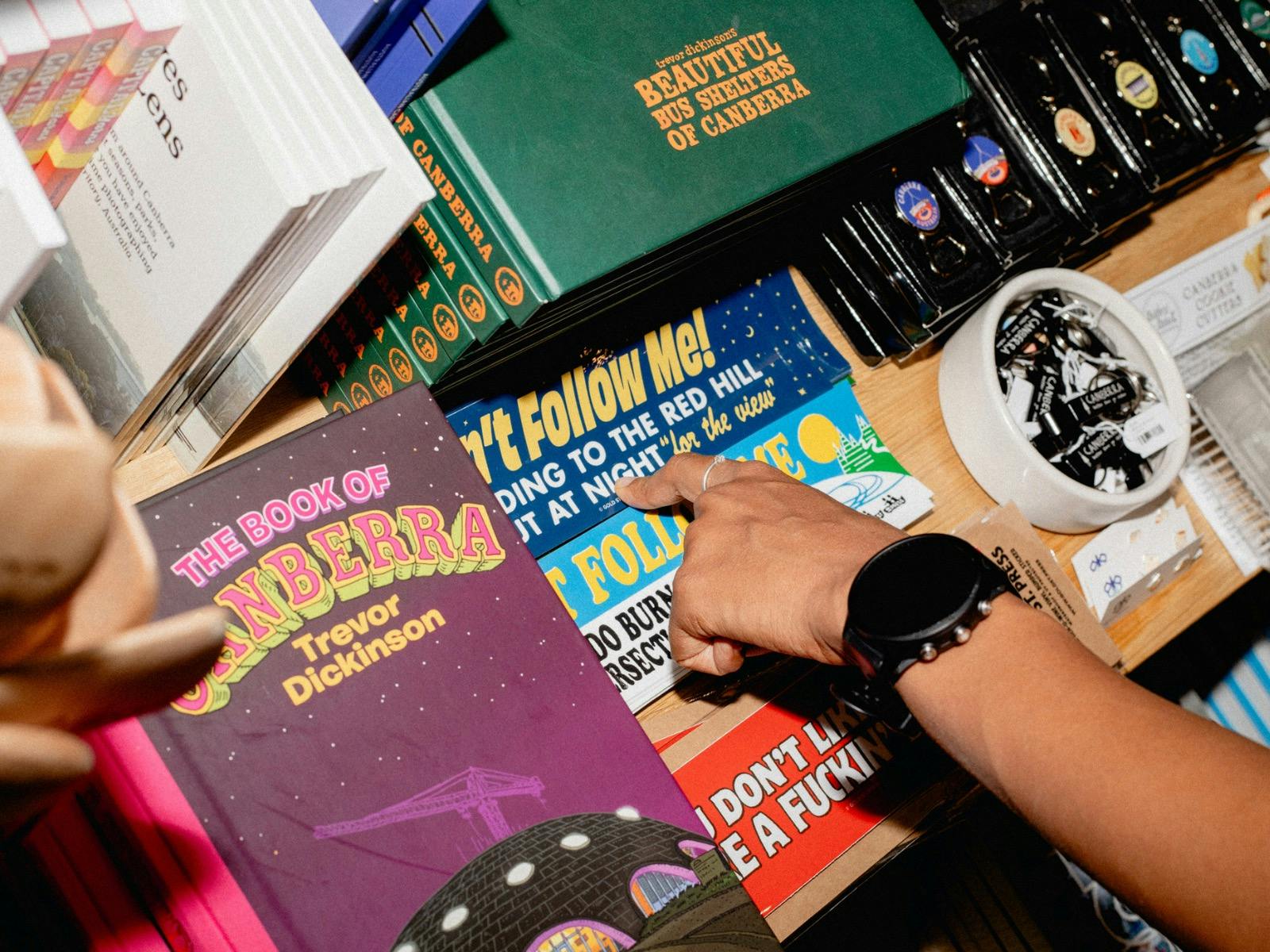 A close-up of a shopper’s hand browsing a selection of Canberra-themed books, stickers