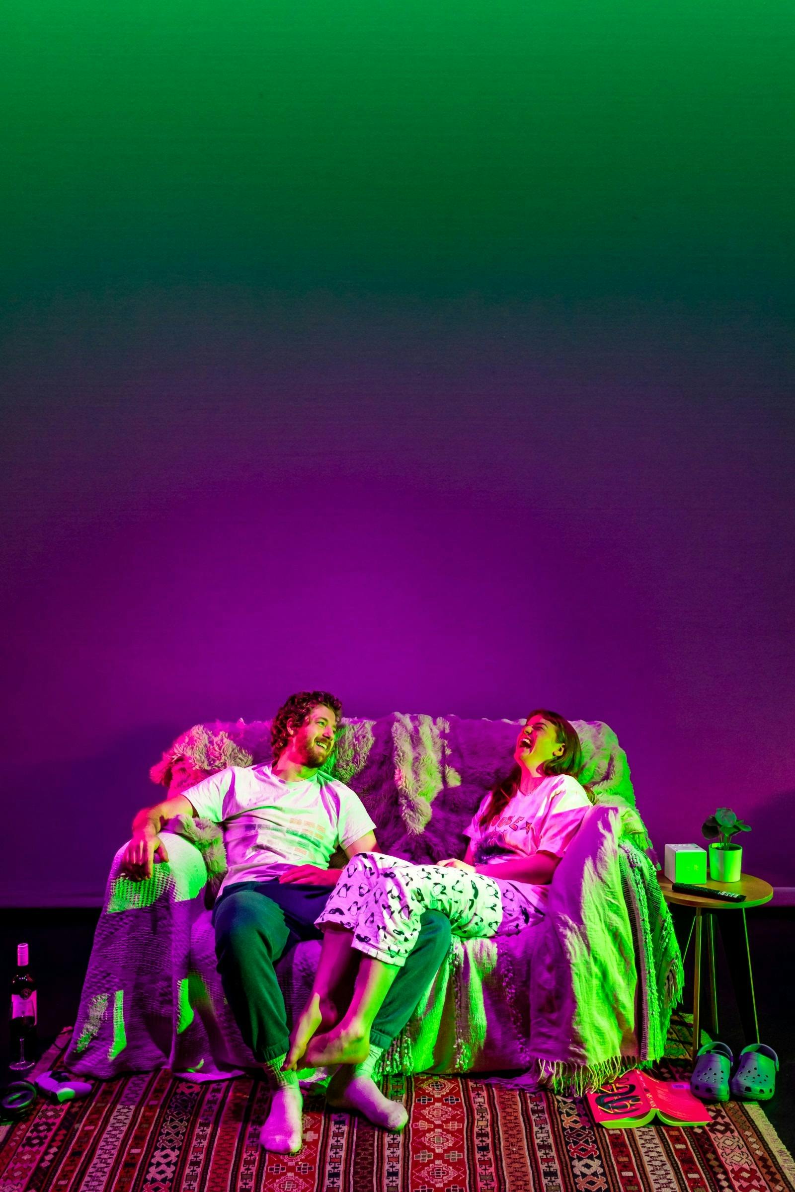 A couple laughs together on a sofa under vibrant green and purple lighting.