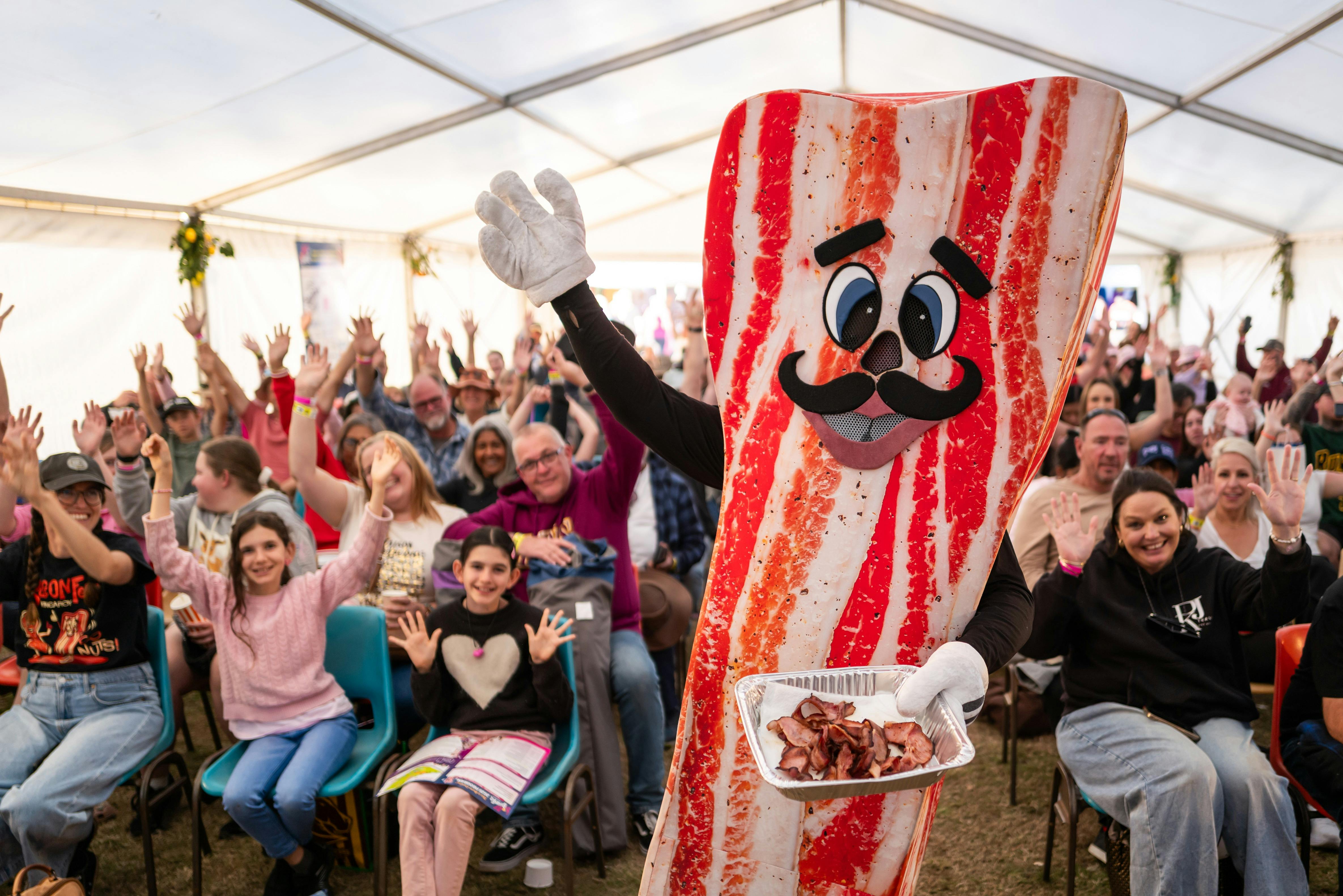 Festival mascot BaconMan is holding a tray of bacon and standing in front of a crowd of people.