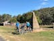 Three riders inspecting tall concrete pyramid shaped monument on roadside grassland