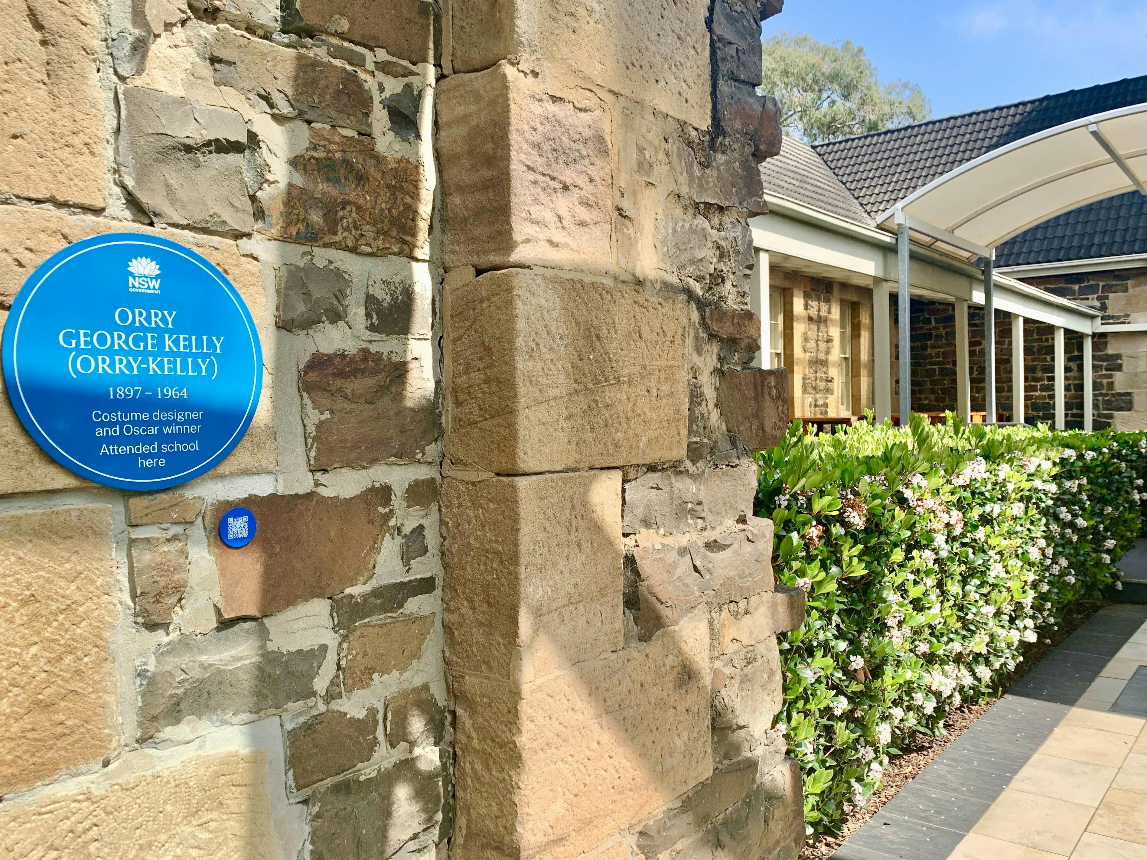 Blue plaque on sandstone brick wall along garden lined pathway