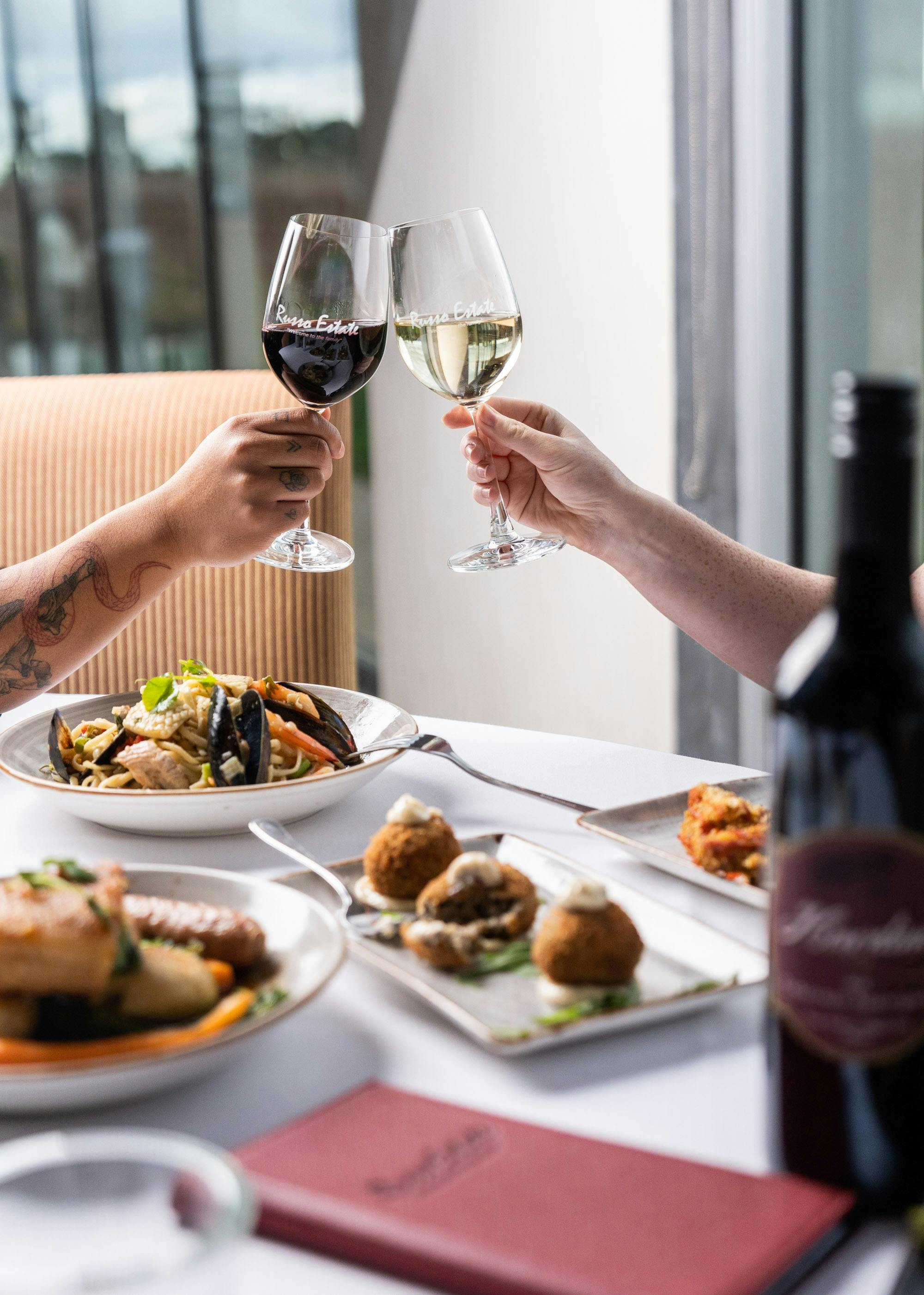 Two people clinking wine glasses in a 'cheers' with an array of our food on the table