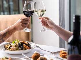 Two people clinking wine glasses in a 'cheers' with an array of our food on the table