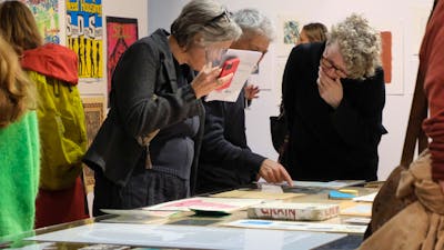 Three people look at artworks on paper upon a long table