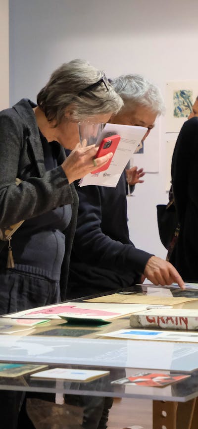 Three people look at artworks on paper upon a long table