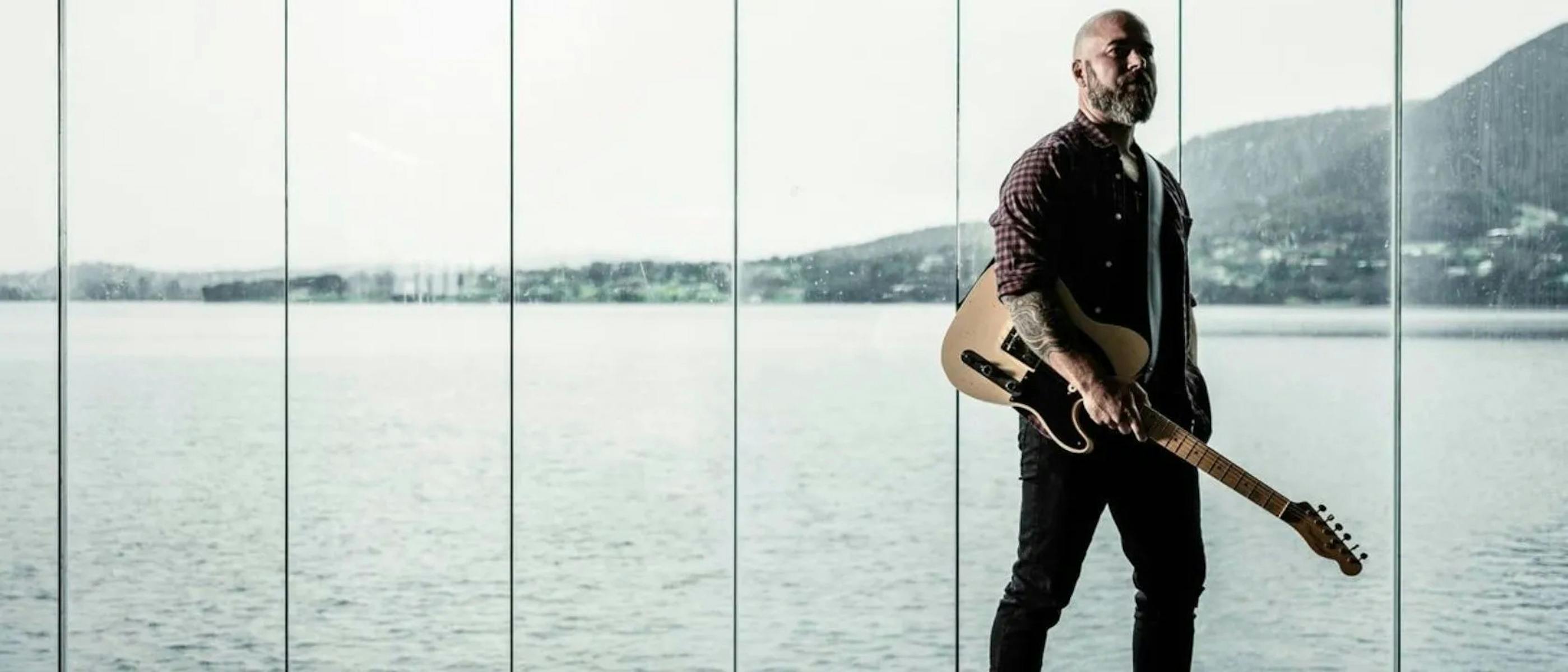 A man holding a guitar stands in front of a window, with water and hills behind him.