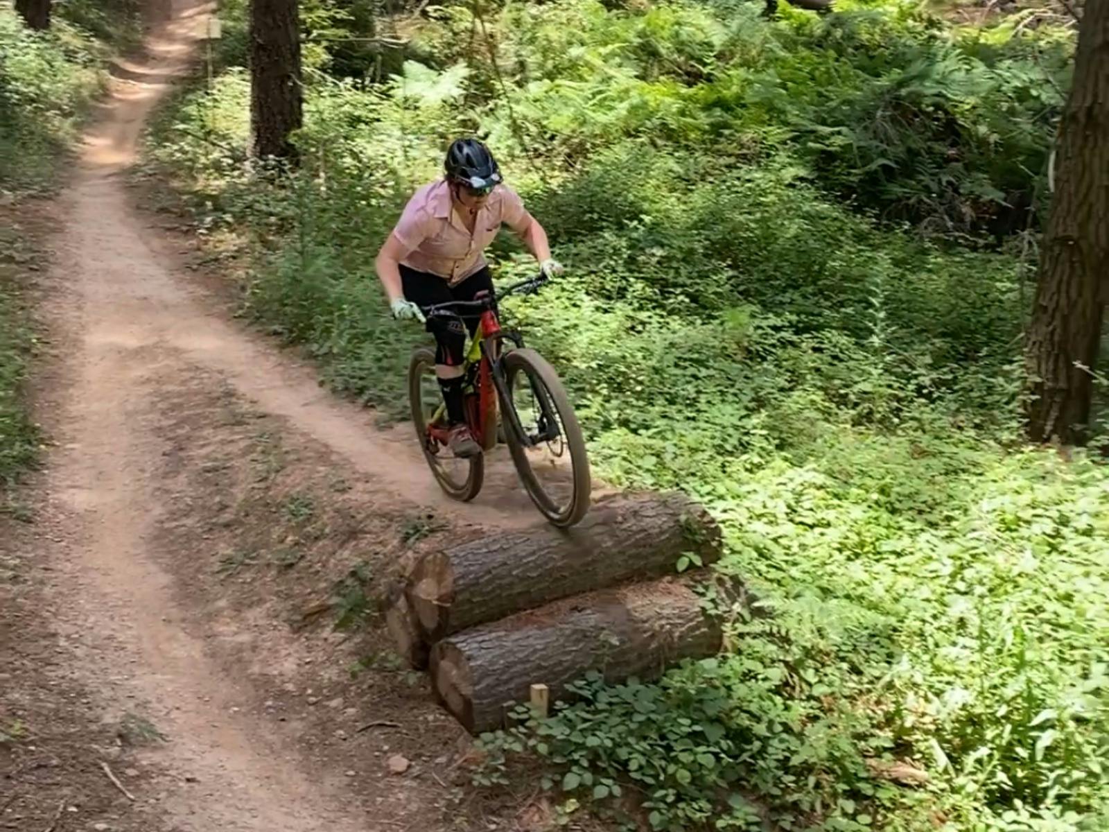 A rider wearing a pink shirt approaches a jump made of logs on a trail.