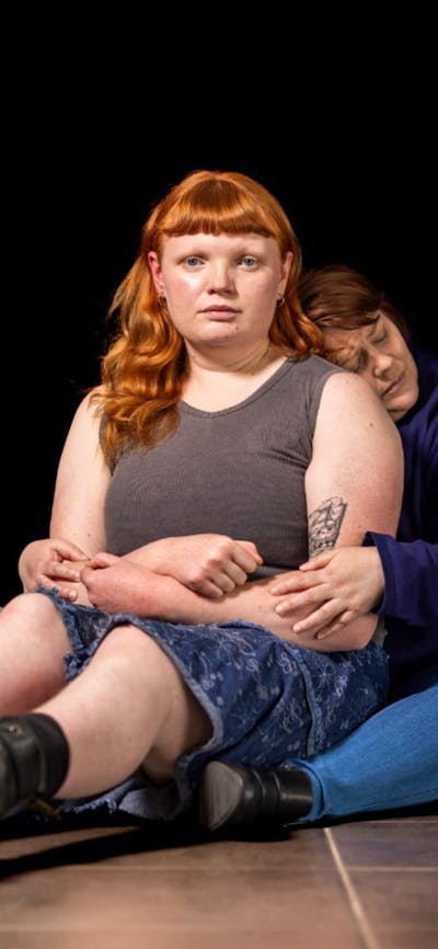 A girl sits on the floor with her legs out staring at the camera while a woman holds her in her arms