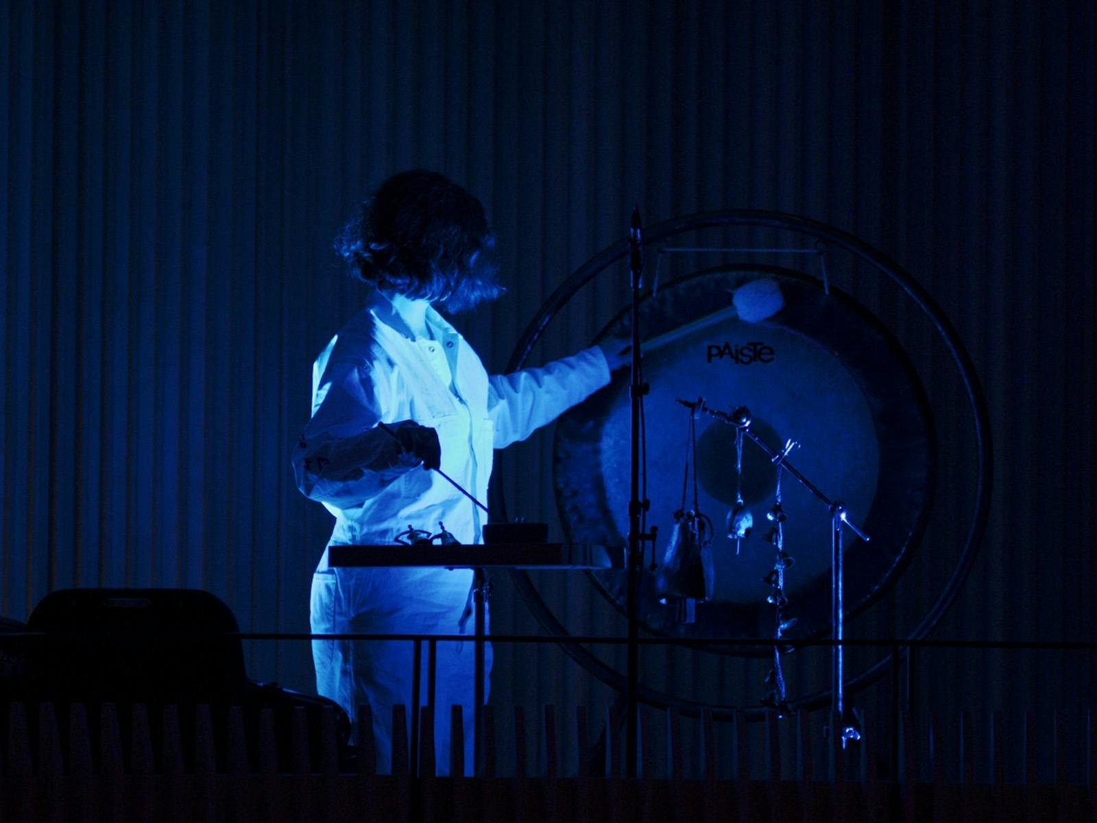 A musician is illuminated softly by a blue light, she is standing playing percussive instruments