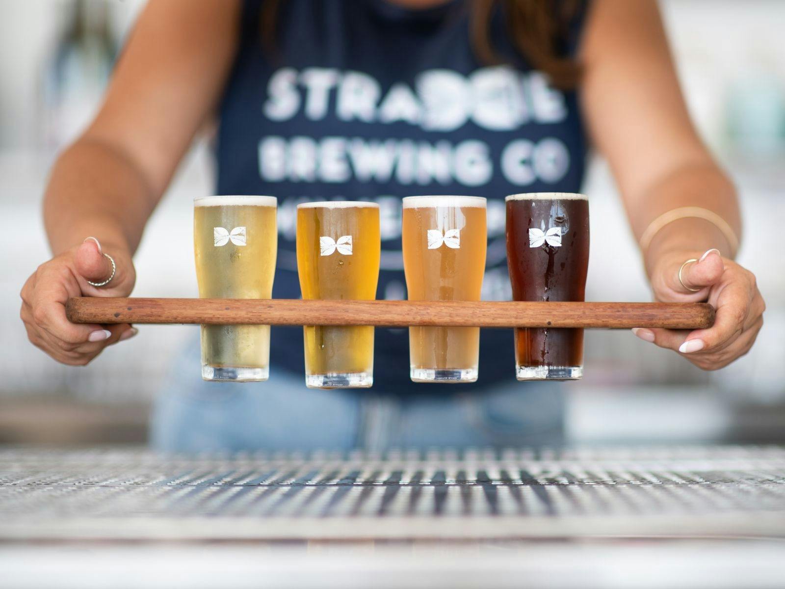 Wooden Paddle containing 4 beers is held out in front of figure in blue shirt