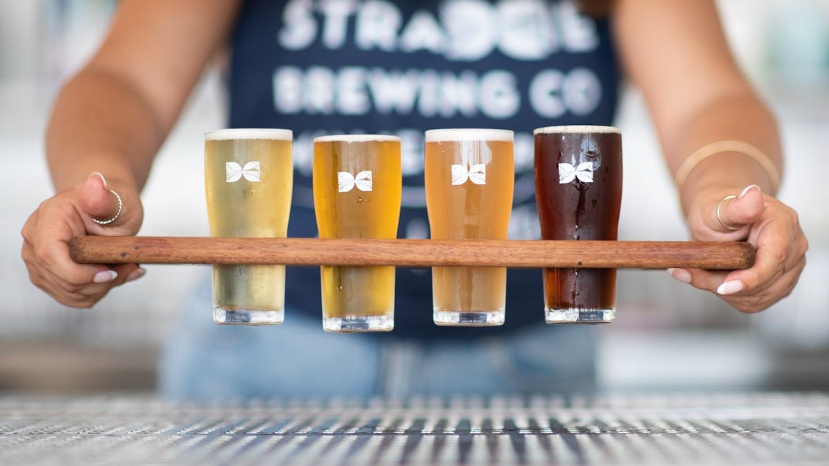 Wooden Paddle containing 4 beers is held out in front of figure in blue shirt