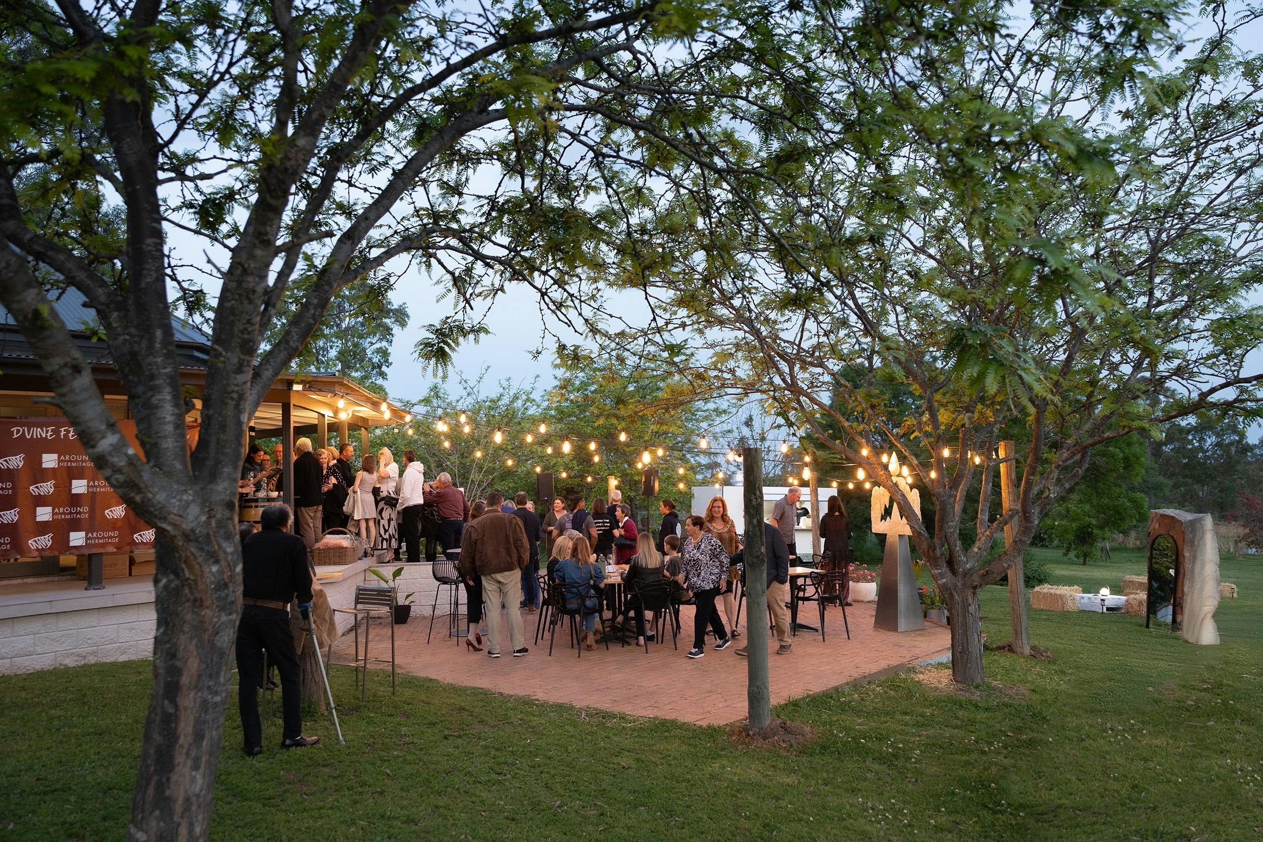 People eat and talk, live music is playing and fairy lights hang between the trees outdoors