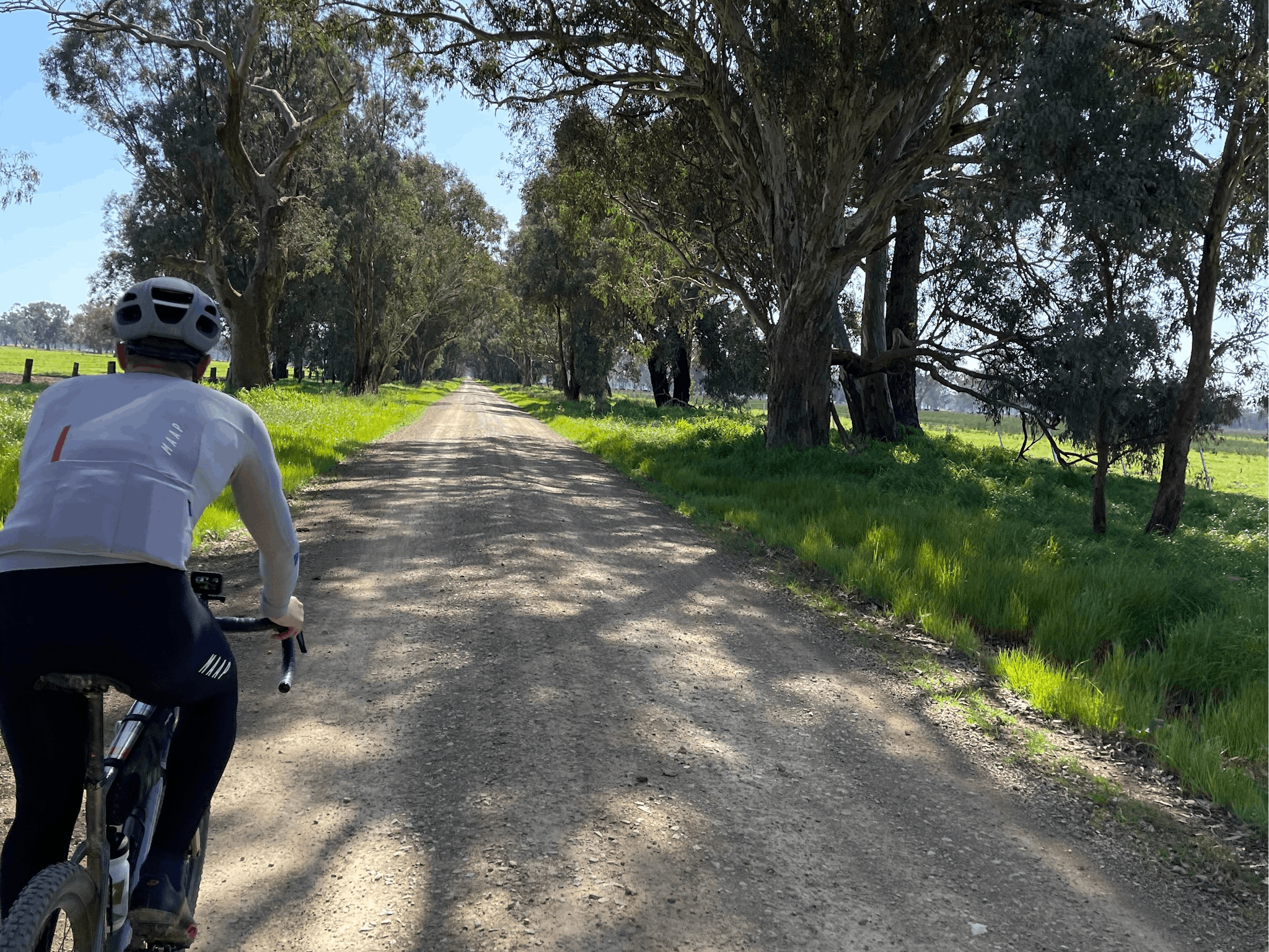 Cyclist on dull brown gravel road, grass, gum trees, blue sky