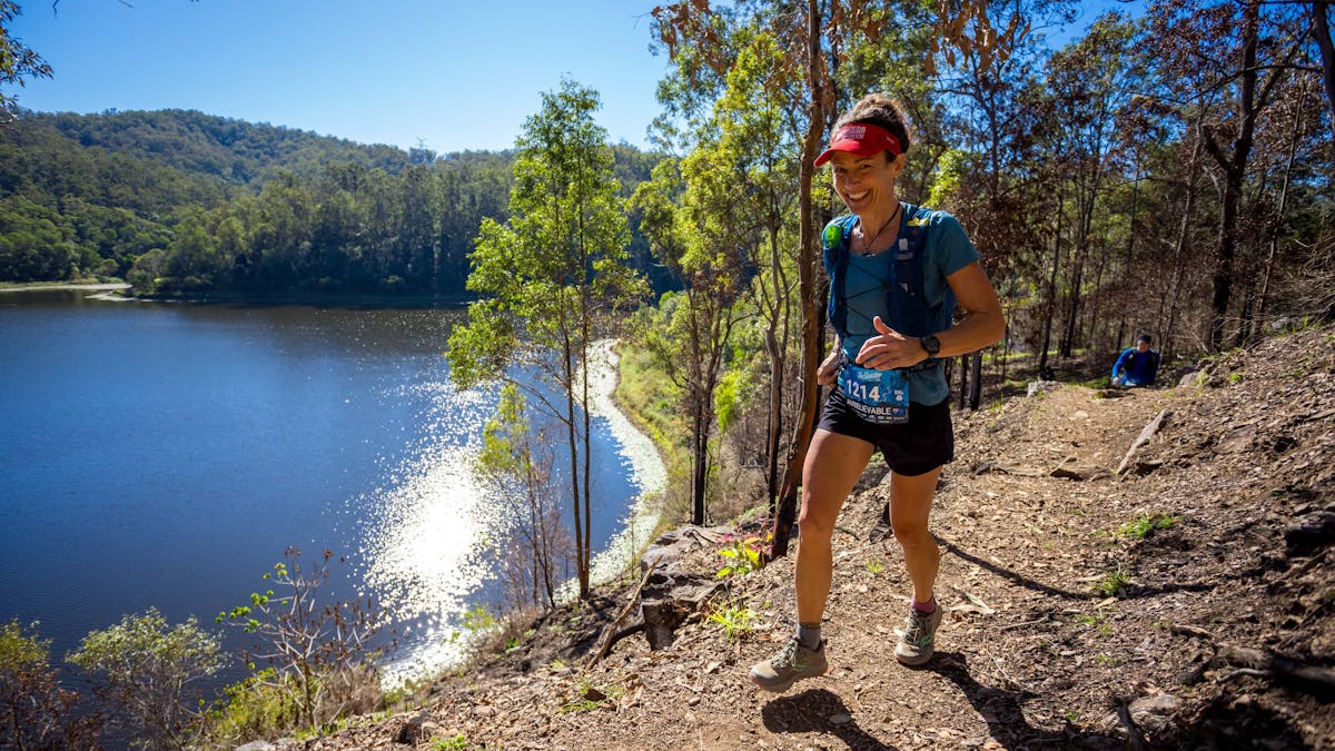 A smiling runner passes on the trail alongside a lake of water