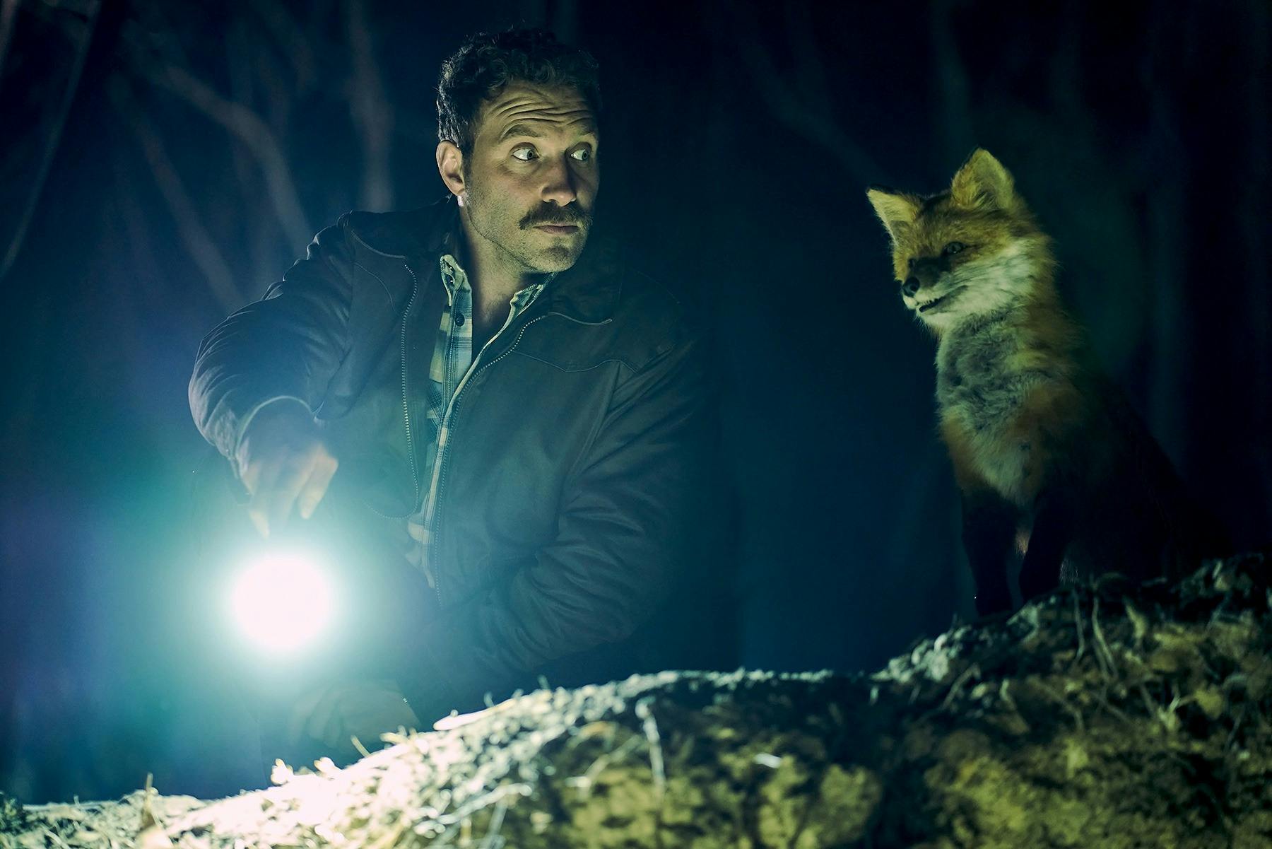 A man shines a torch whilst sitting next to a fox.