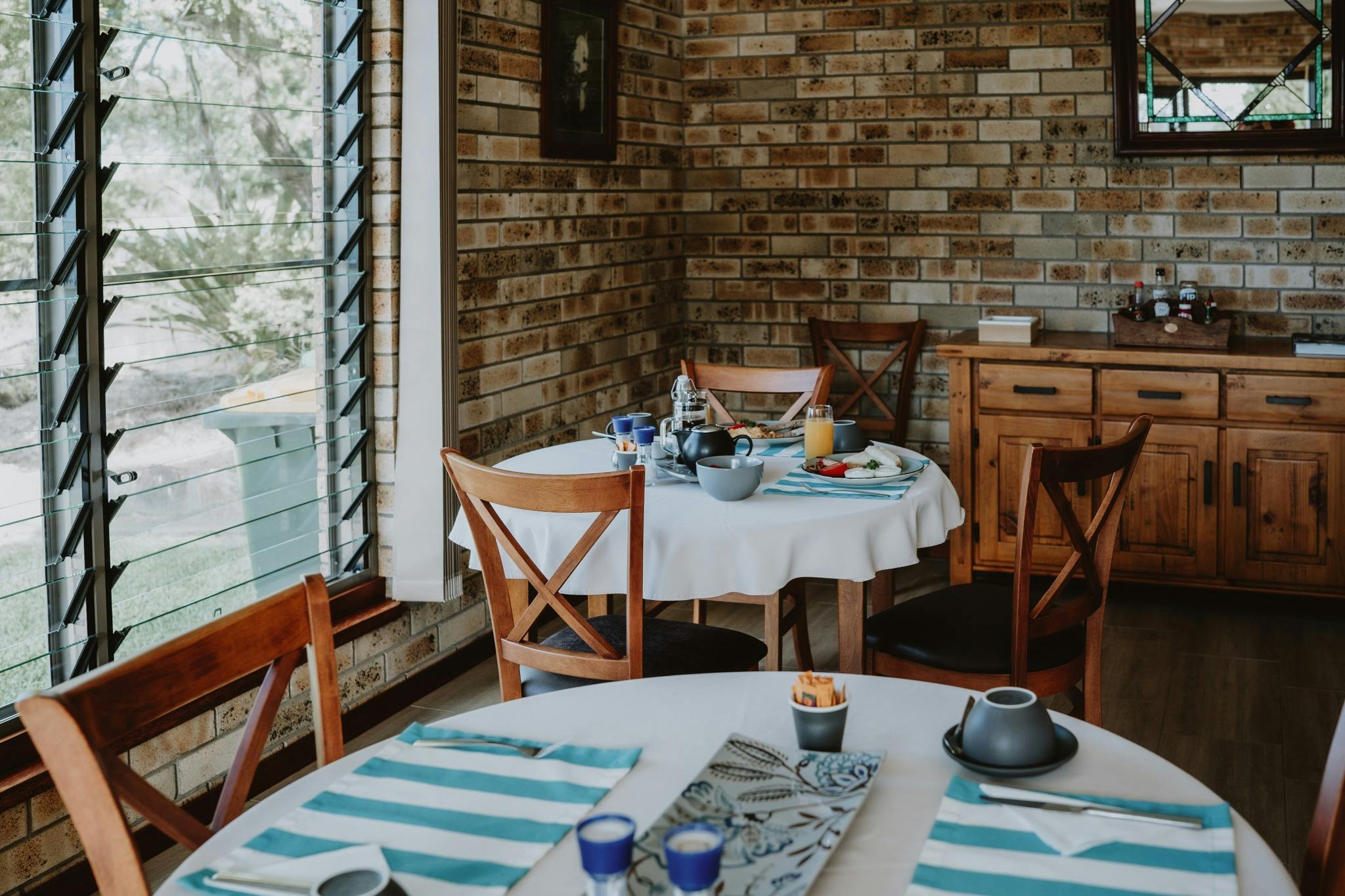 Common area dining room with table settings, by louvre windows with hutch in the background