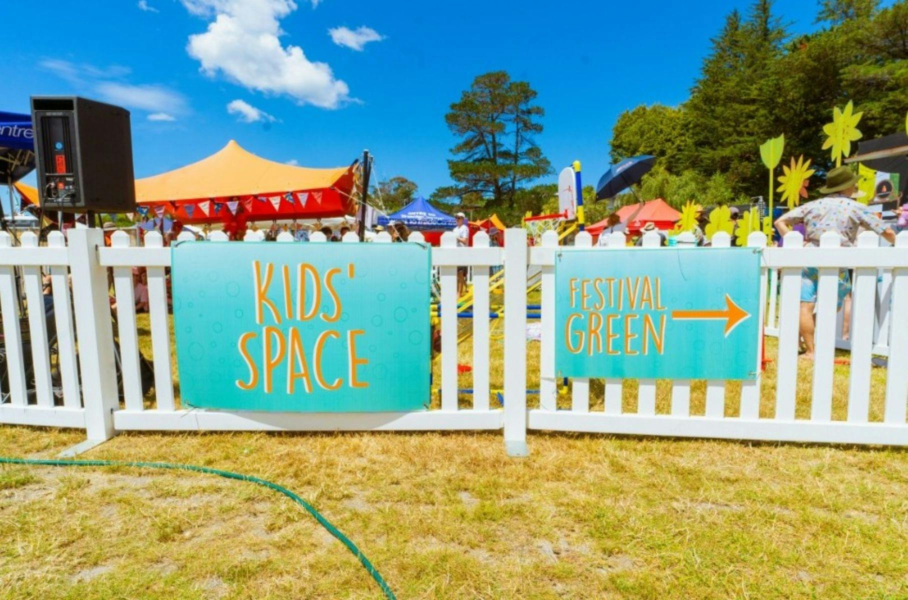 White picket fence with two signs reading Kids Space and Festival Green