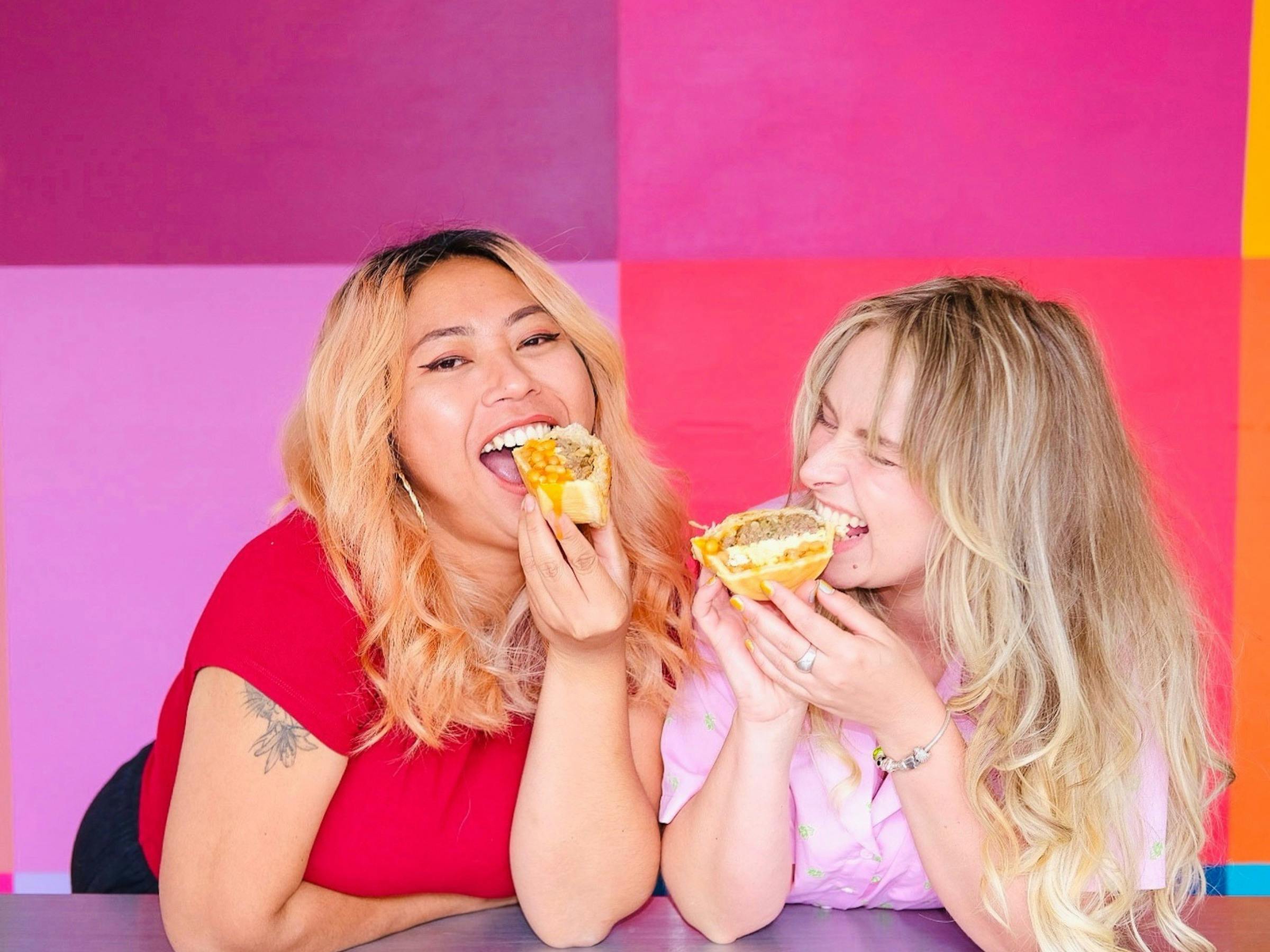 Two people smiling and biting into a pie in front of a colourful backdrop