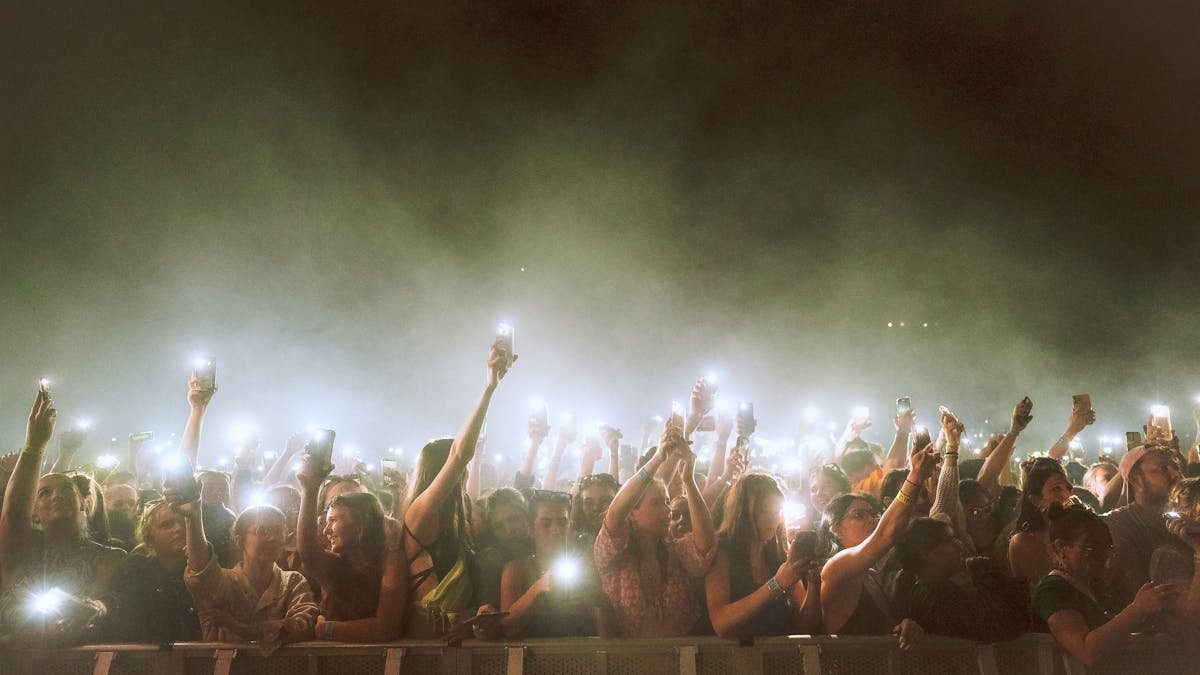 A large crowd stands behind a barrier holding their phones with their torch light on.