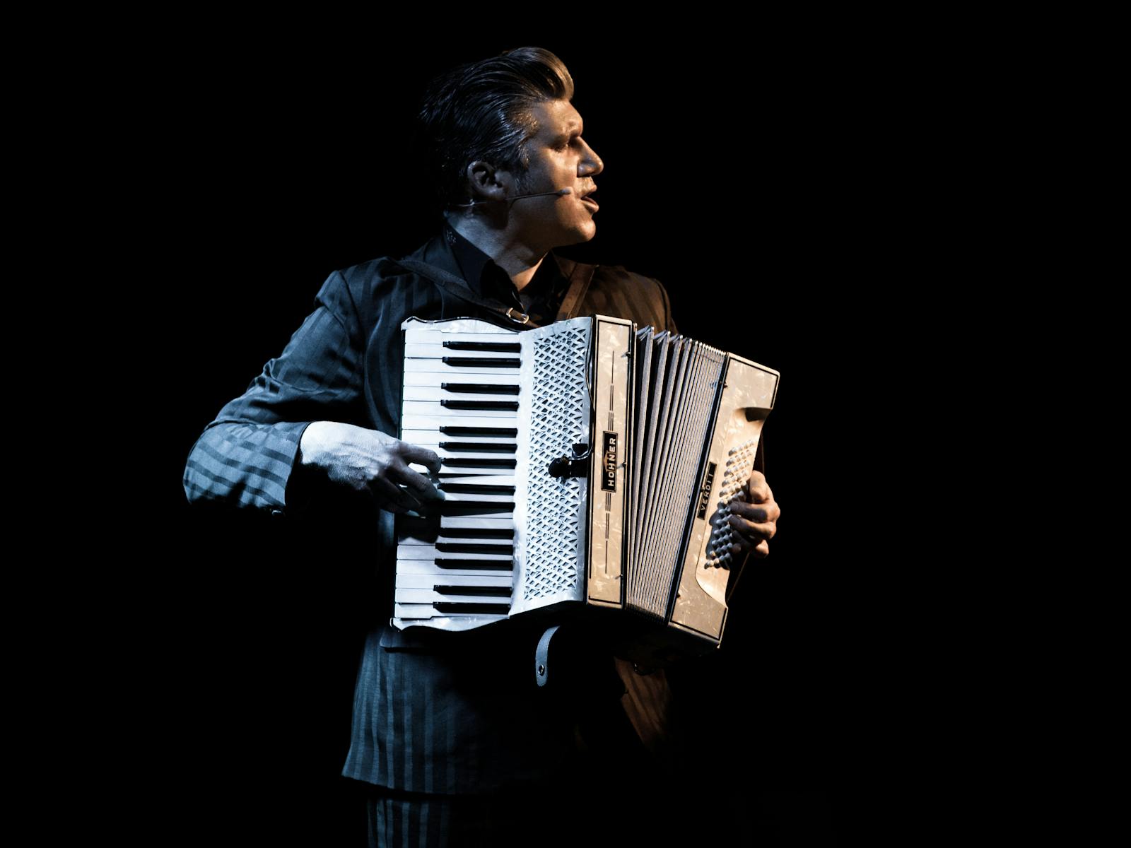 A man looks to the right holding a piano accordion.  He is lit like he's on stage.