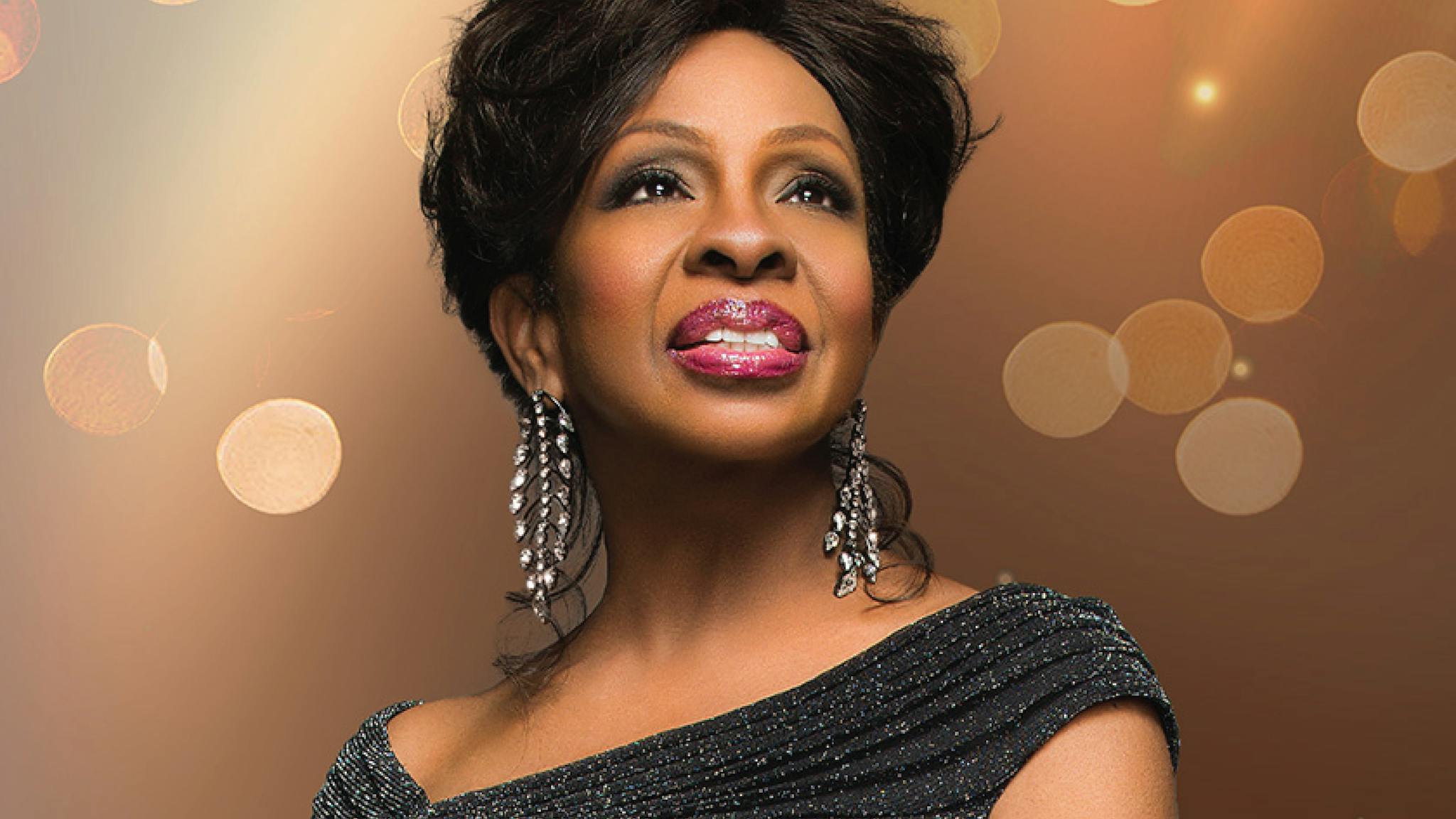 Gladys Knight in a black dress looking to the right in front of a glittery brown backdrop