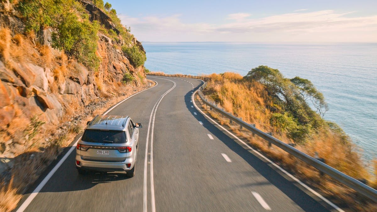 A vehicle drives uphill along a winding road, leaving the beach below in the distance