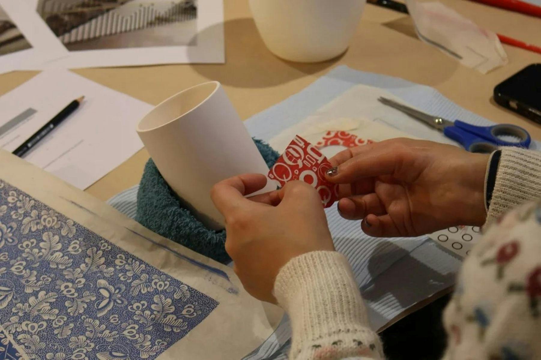 A close up shot of a pair of hands applying a red decal to a white  ceramic cup