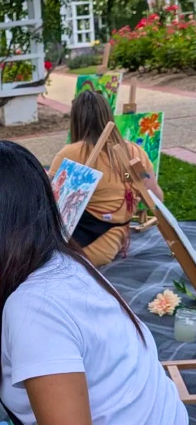 A woman seated on the ground in a rose garden paints a colourful bouquet of flowers on a canvas.
