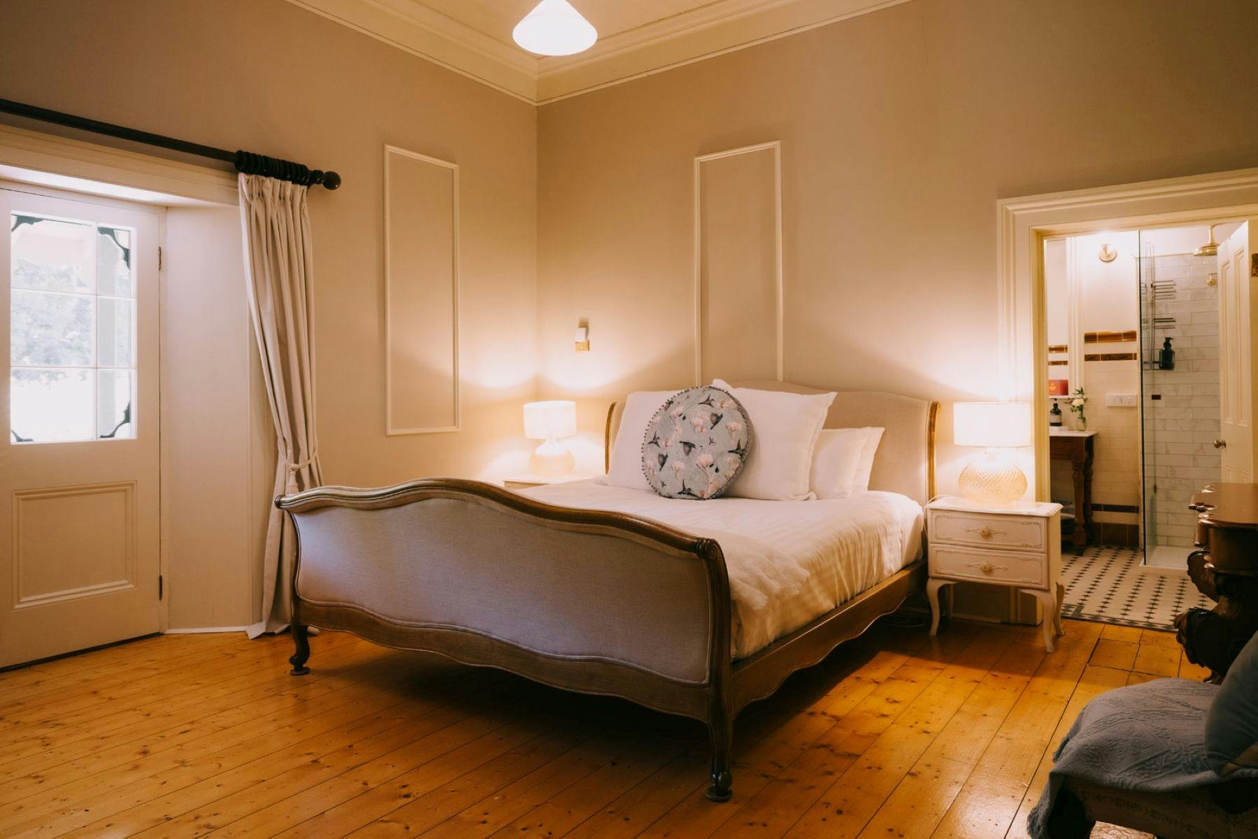 A bedroom featuring a vintage-style bed, warm lighting, wooden floors, and an ensuite bathroom.