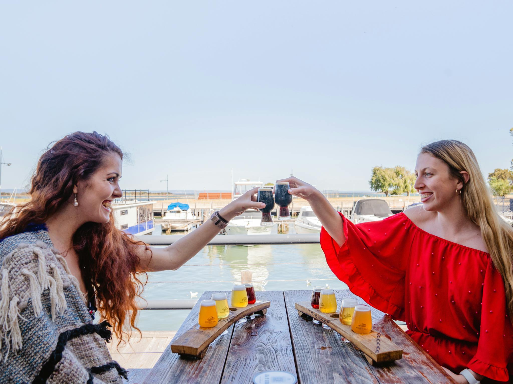 Two girls cheersing their beers on an outdoor deck overlooking the water at Boundary Island Brewery