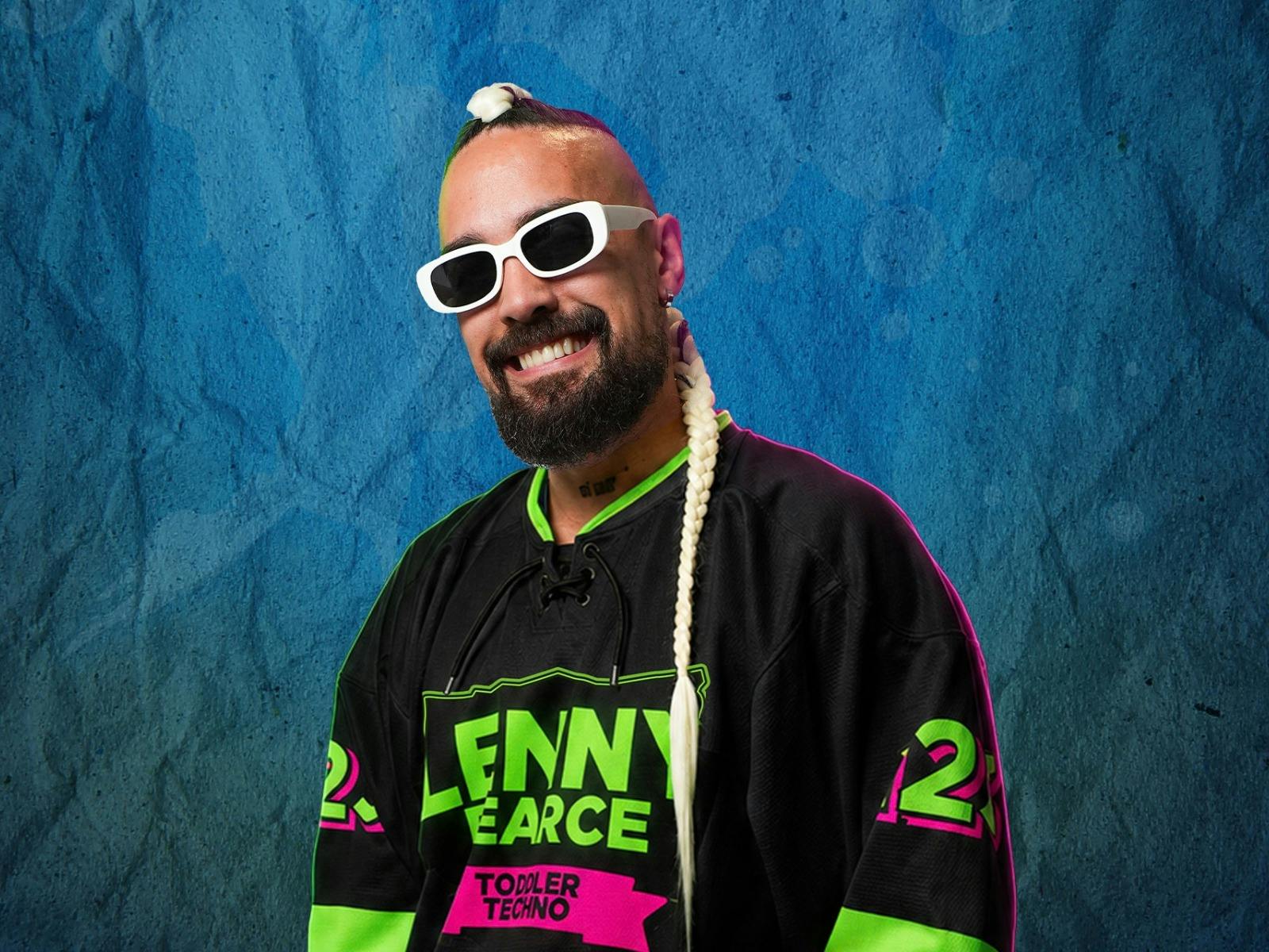 A smiling man wearing white sunglasses, a long blonde braid, and a black jersey poses.