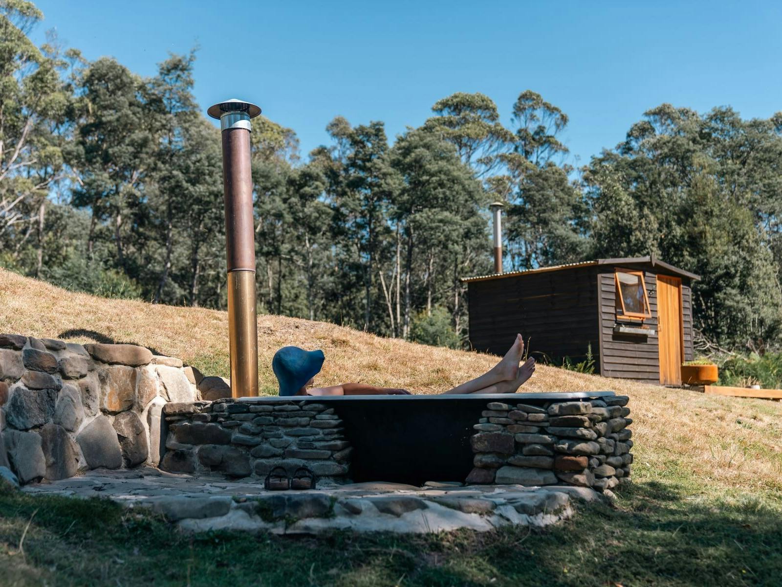 A figure in an outdoor bath tub with a rustic cabin and forest in background.