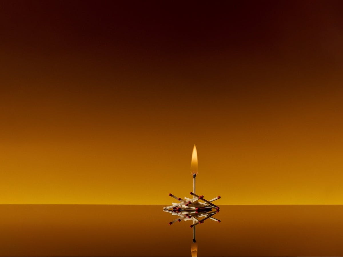 A single lit matchstick sitting on top of other unlit matchsticks. Warm Orange background