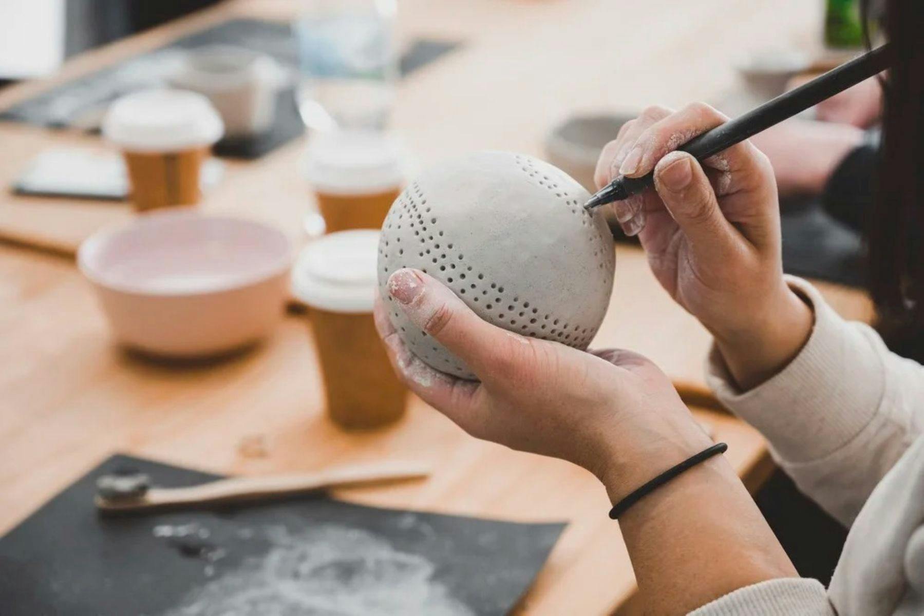 Hands hold a clay sphere, poking holes in with a tool for decoration