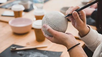 Hands hold a clay sphere, poking holes in with a tool for decoration