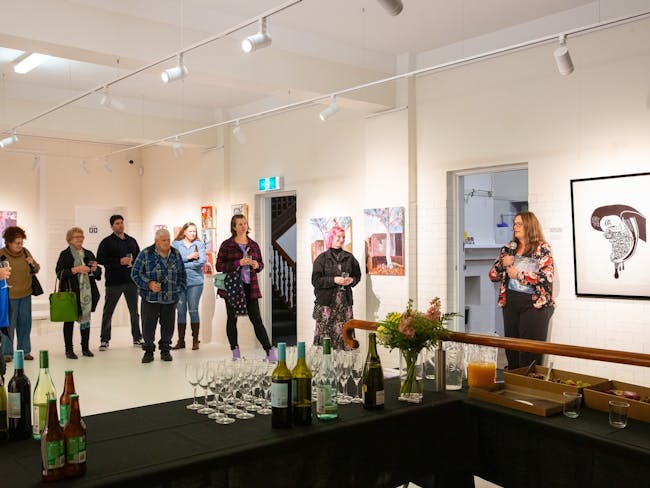 An artist holding a microphone standing in a gallery with people standing around listening