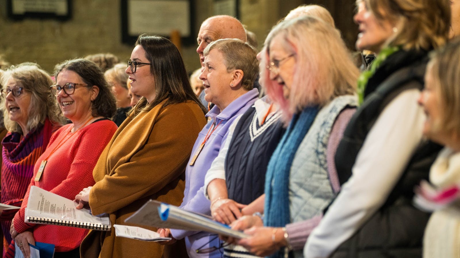 Choir members singing joyfully from scores in a historic, softly lit church setting.