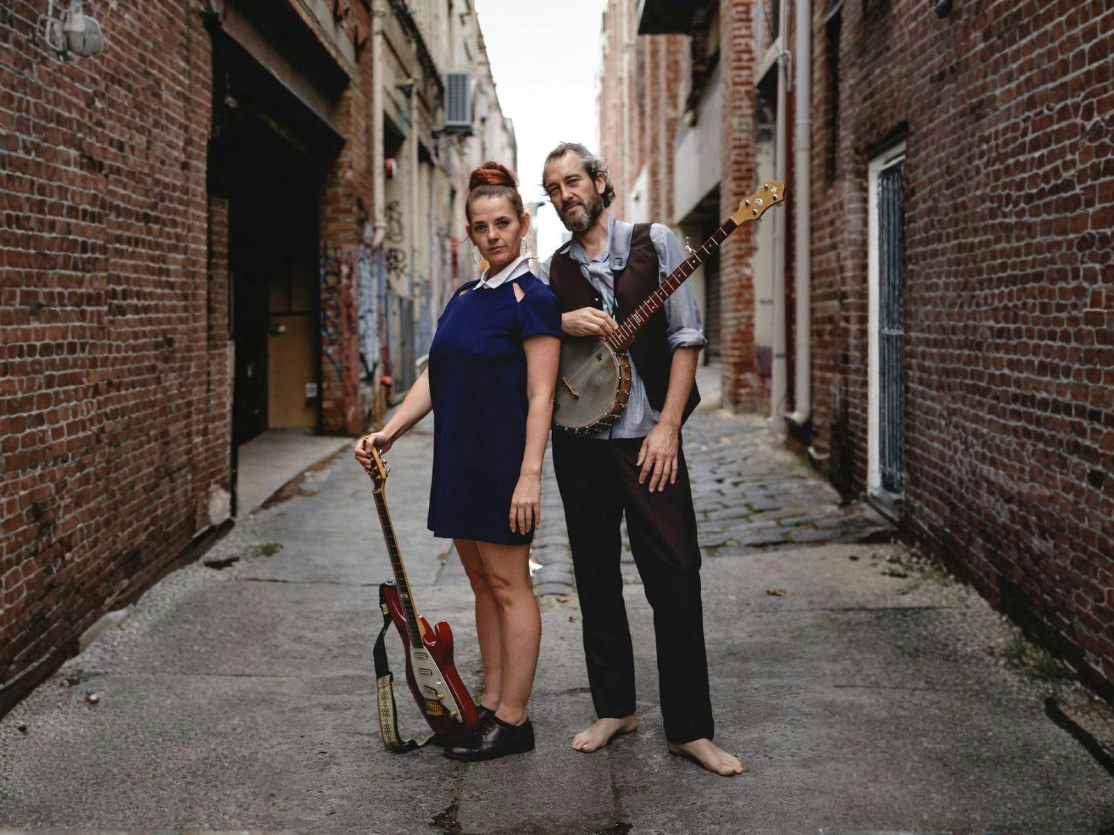Lizzy & Woddy stand in a brick lane holding instruments staring at the camera