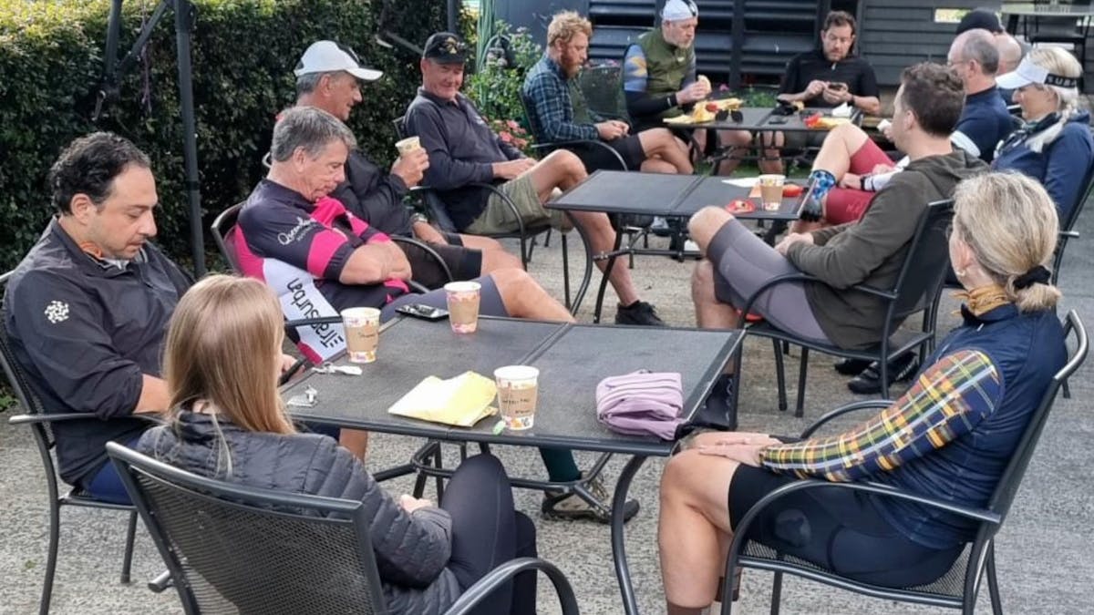A group of cyclists seated at outdoor café tables under shade umbrellas, enjoying drinks .