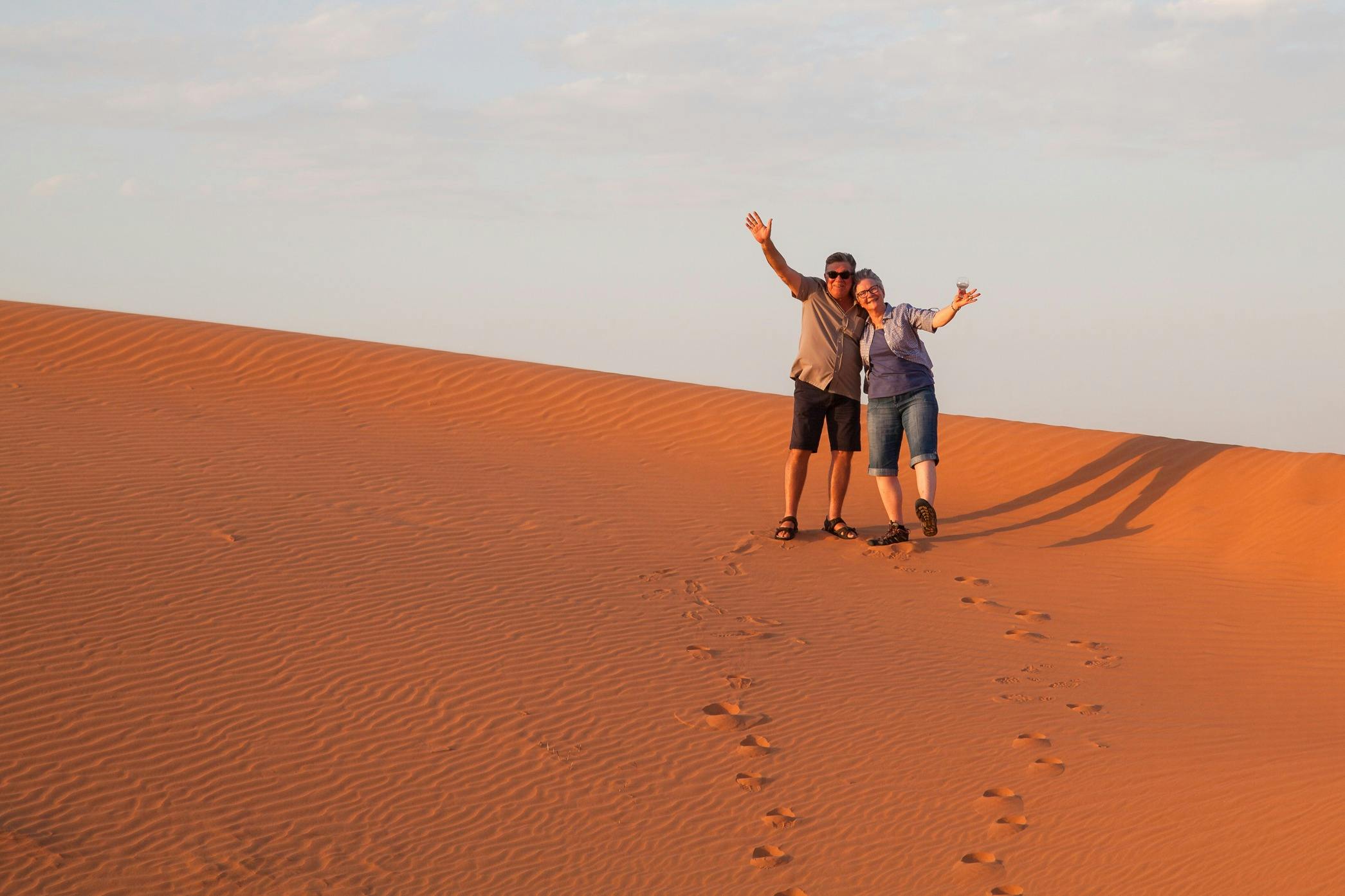A couple waving at the camera while on a big red sand dune