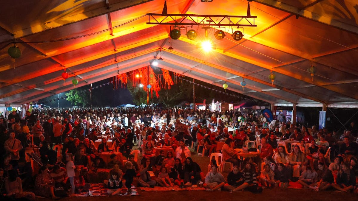 A crowd sits under a large marquee waiting the stage performances.