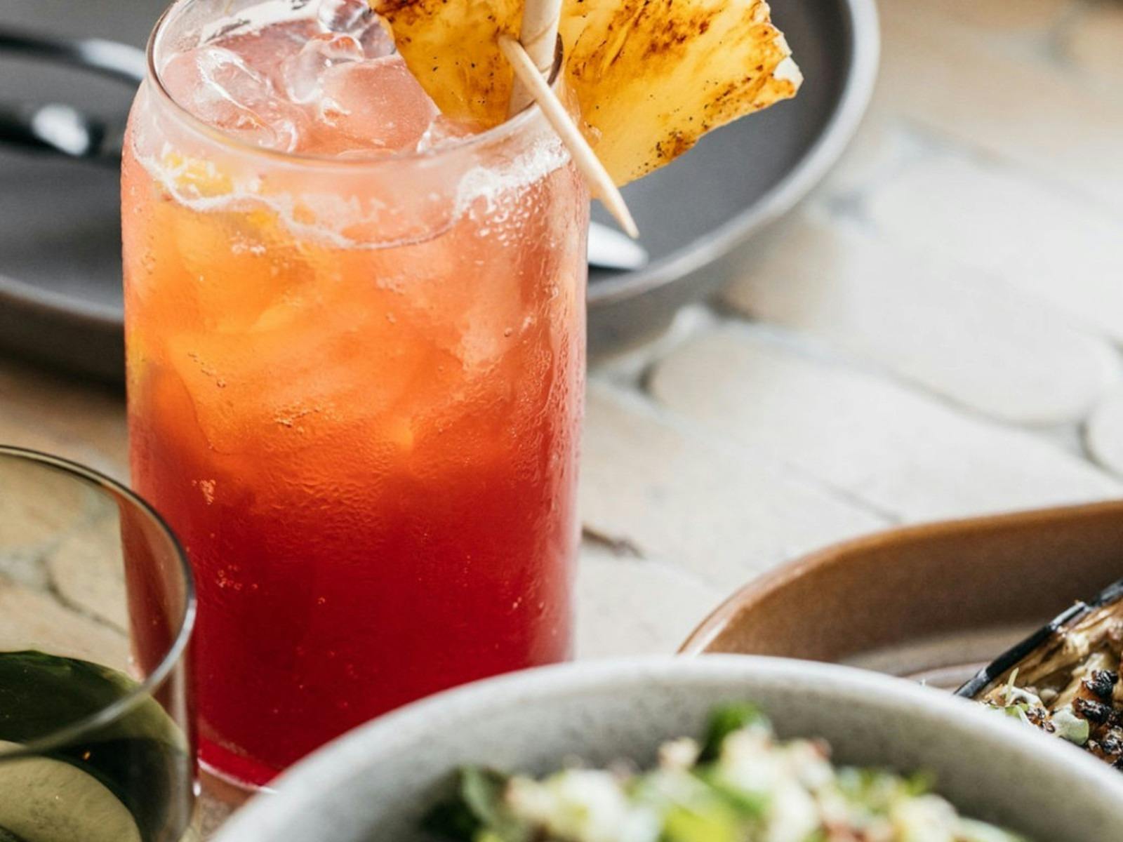 An orange cocktail in a tall glass with a slice of pineapple on top next to plates of food