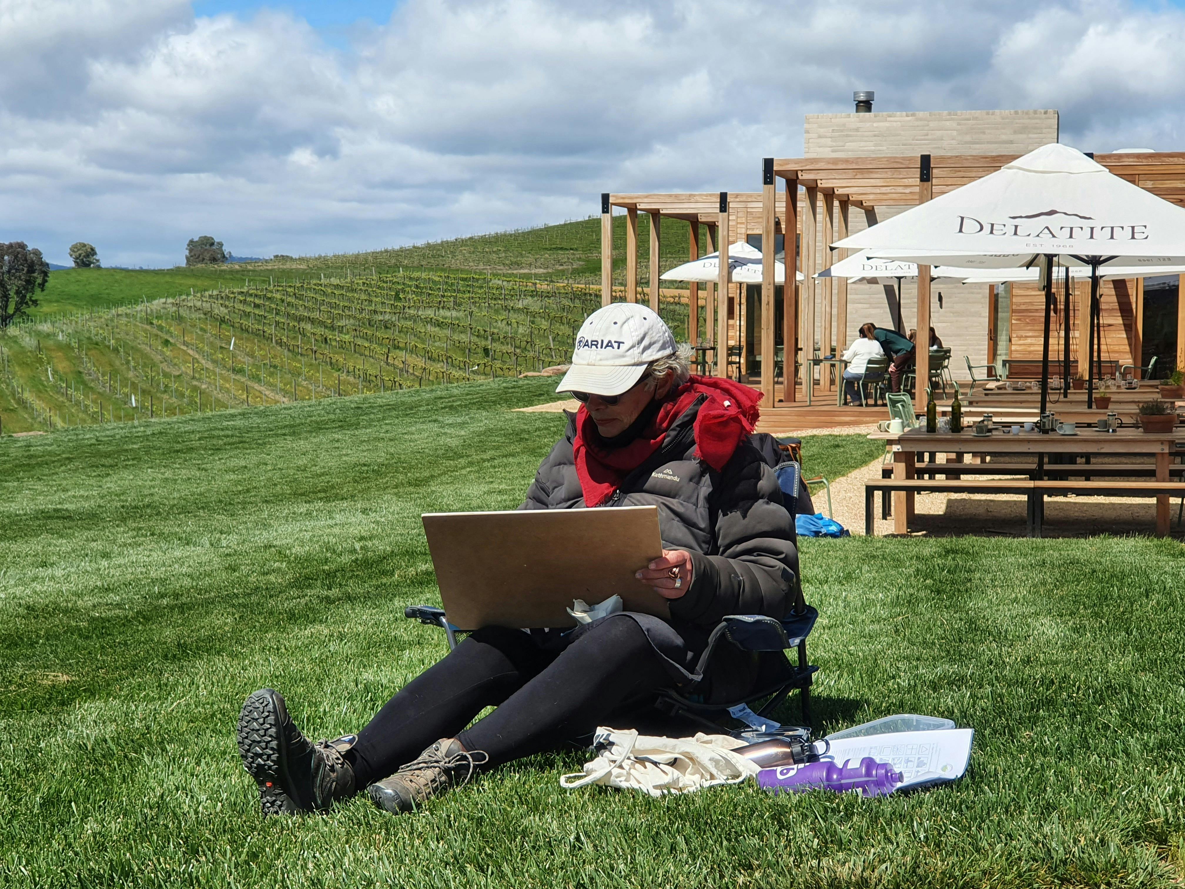 A woman is sitting on lawn in front of winery cellar door, painting.