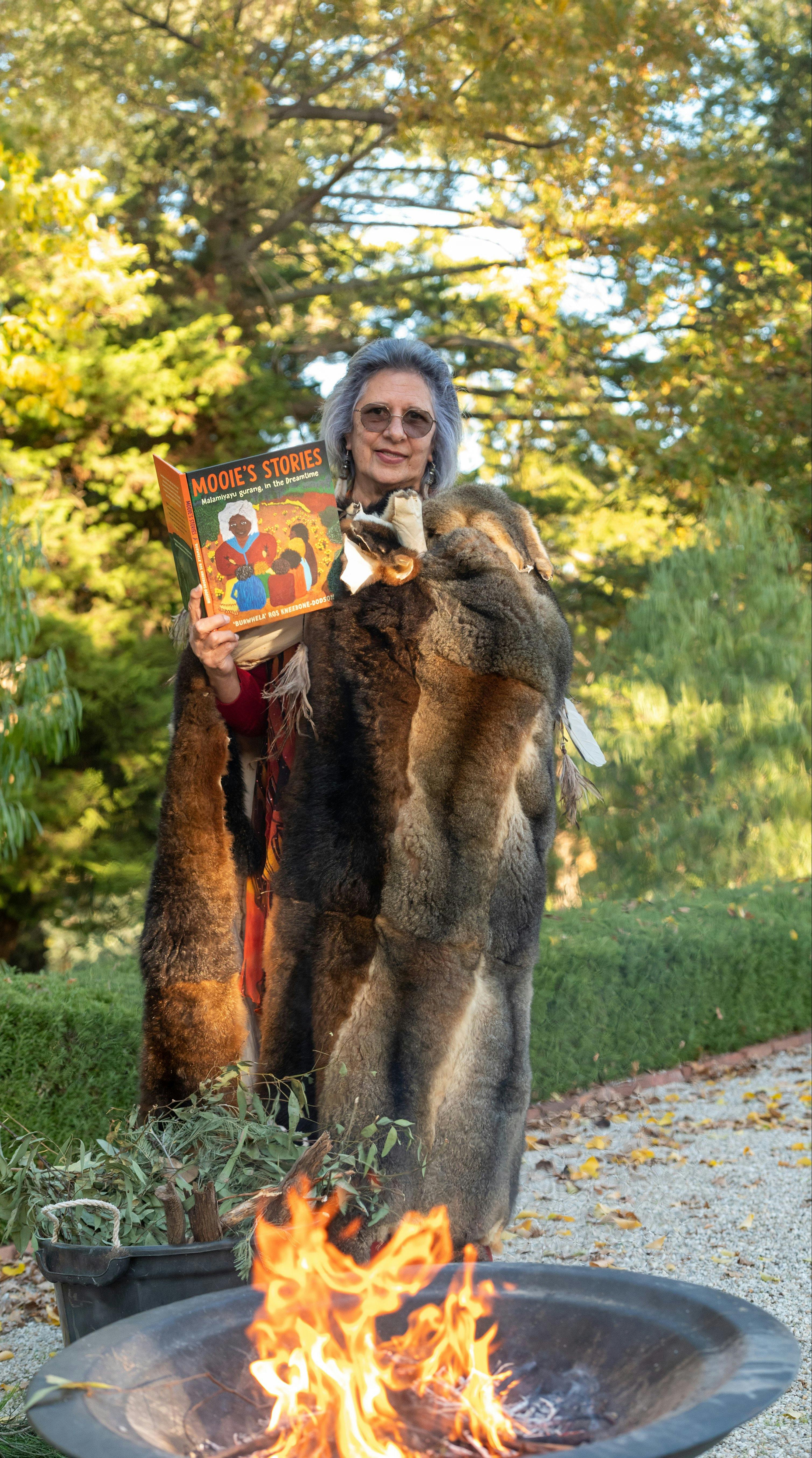 A Djaara woman wearing a possum skin cloak holds a story book
