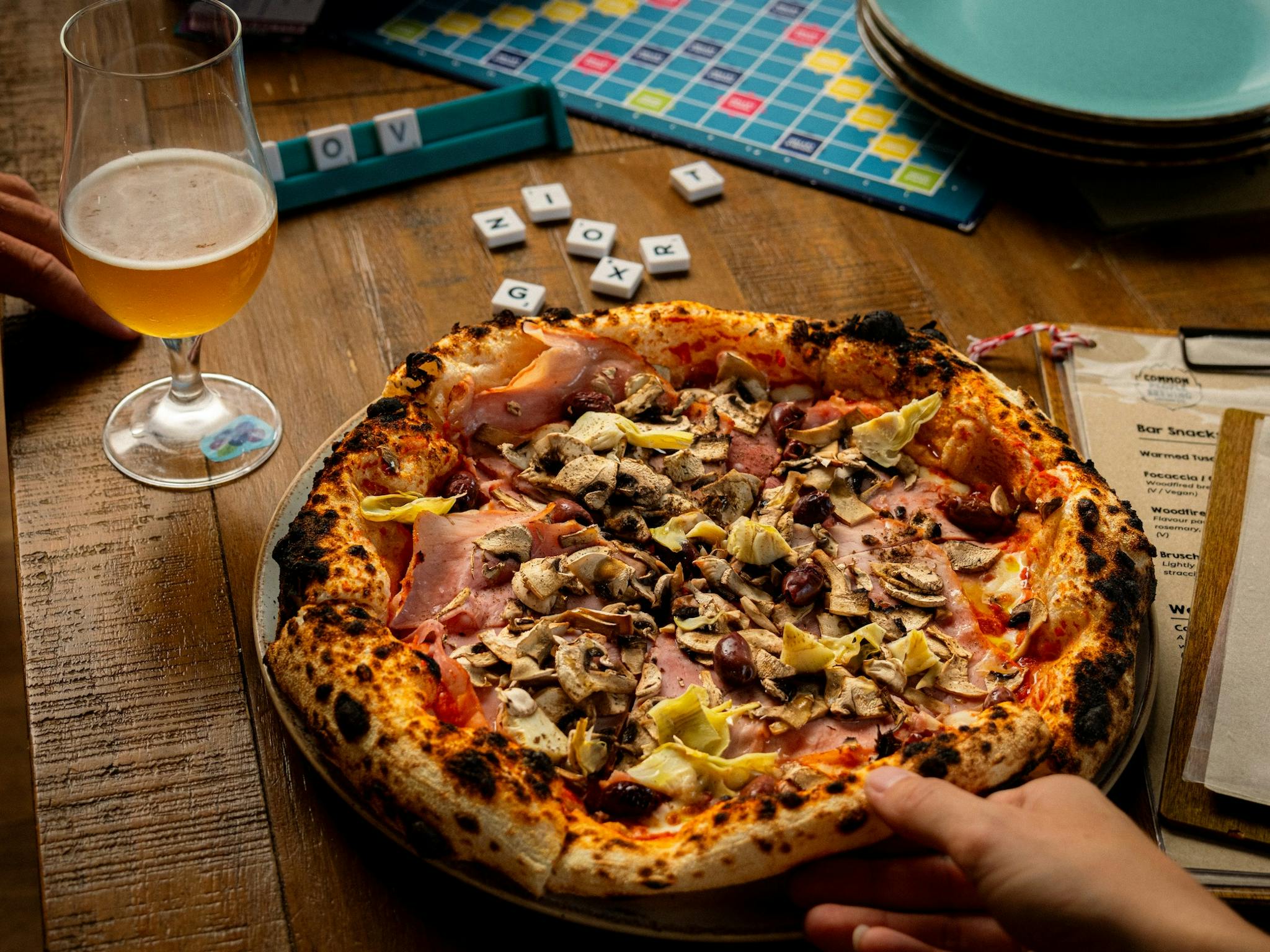 Hand reaching for pizza with a beer, menu and scrabble game pieces