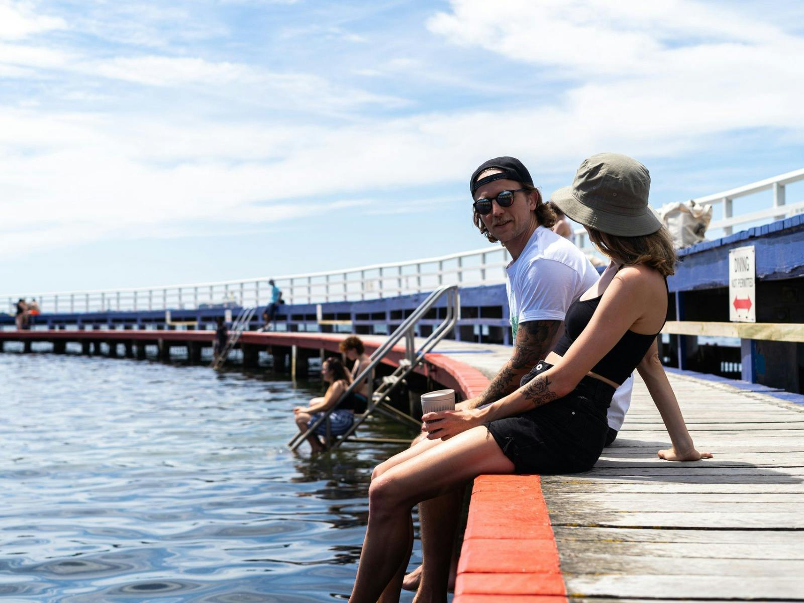 Man and women drinking coffee legs dangling over the wooden walkway of Eastern Beach reserve