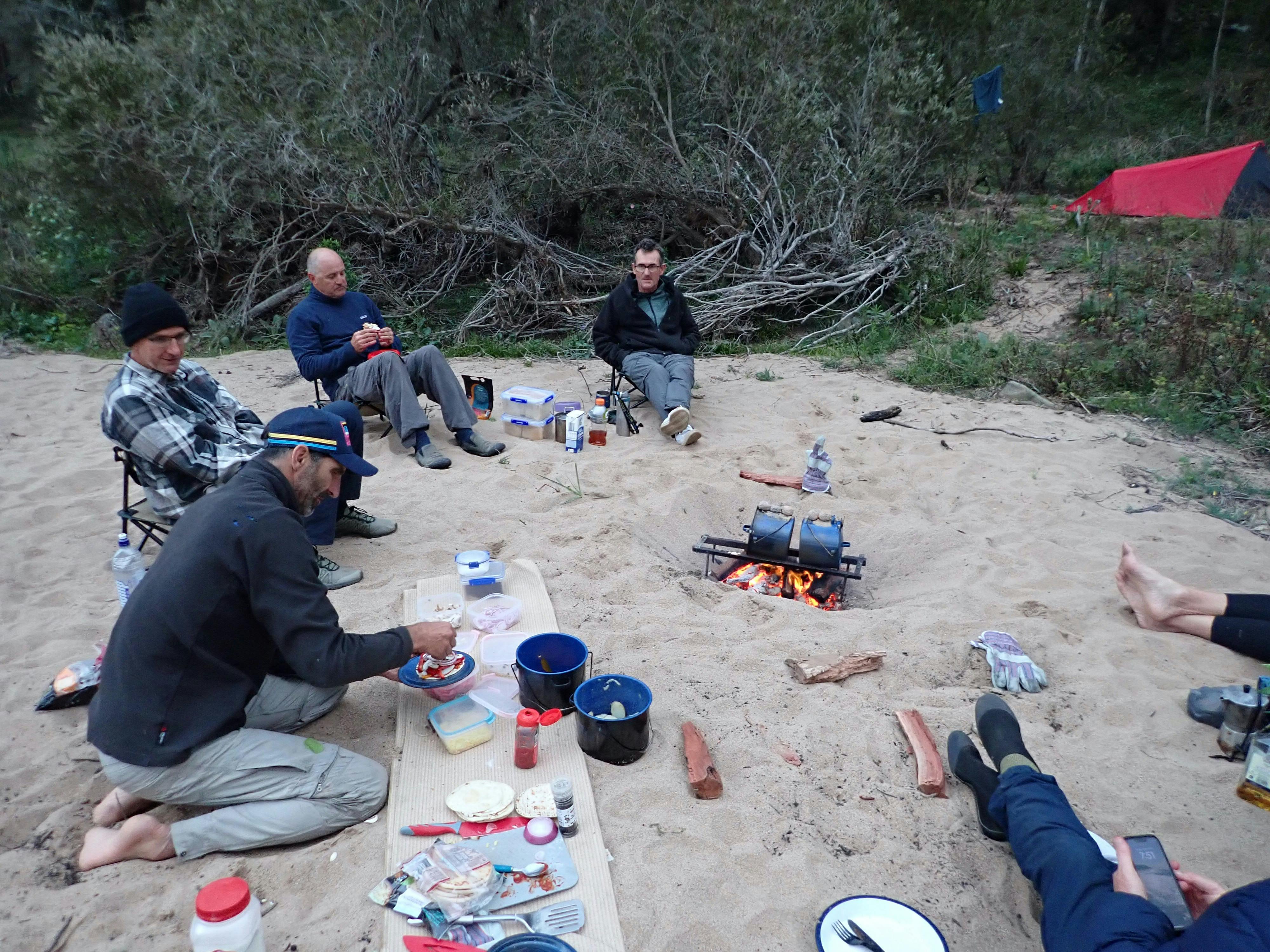Some men are sitting around a fire while a guide cooks dinner on the Snowy River whitewater rafting