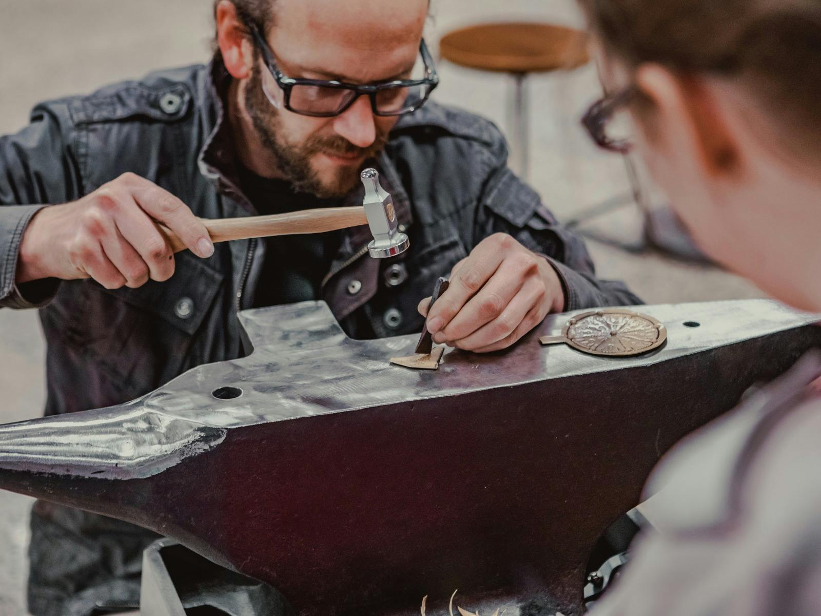 Man hammering metal on anvil while a workshop participant looks on