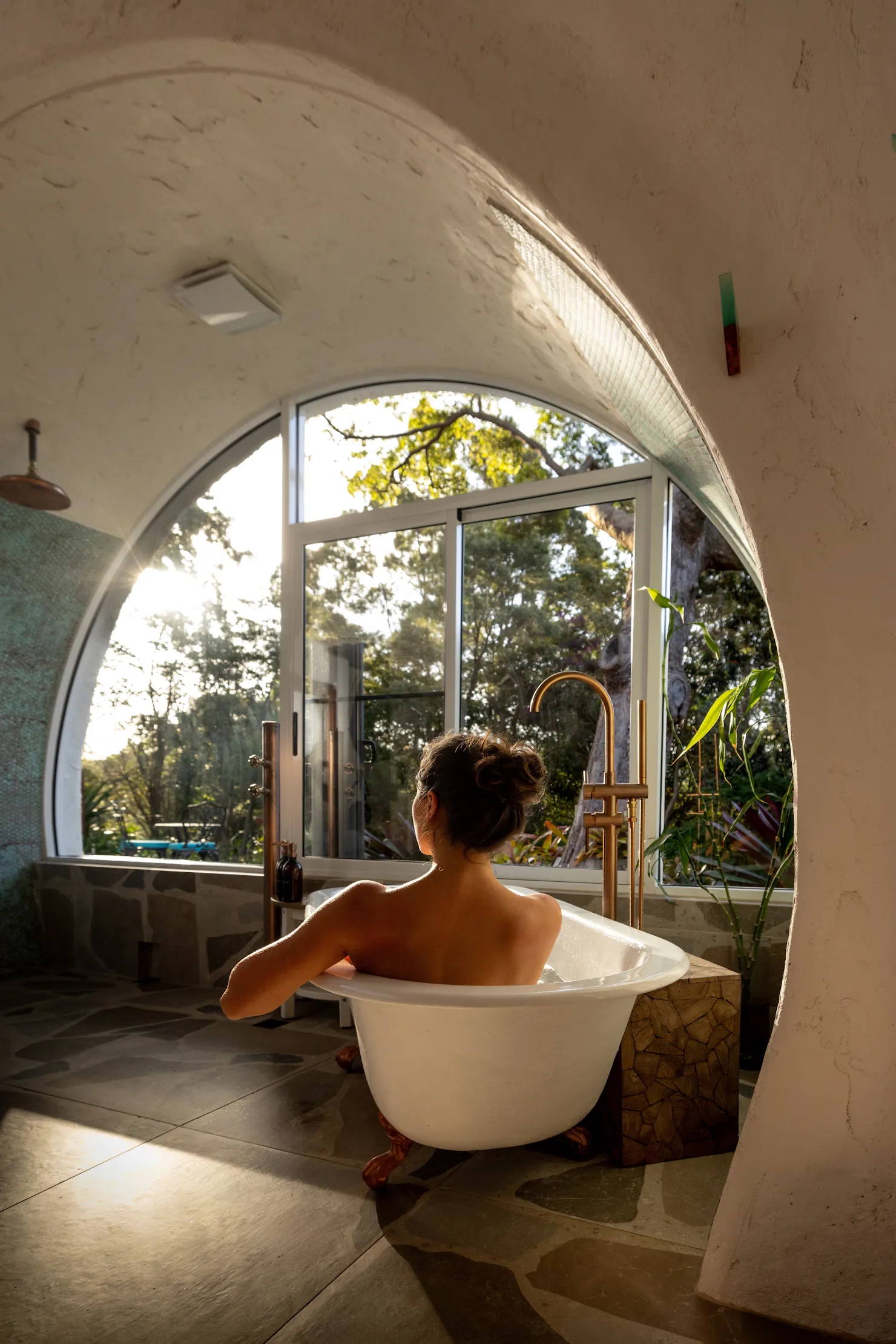 Guest relaxing in a freestanding bath beside a large arched opening overlooking the lush gardens at