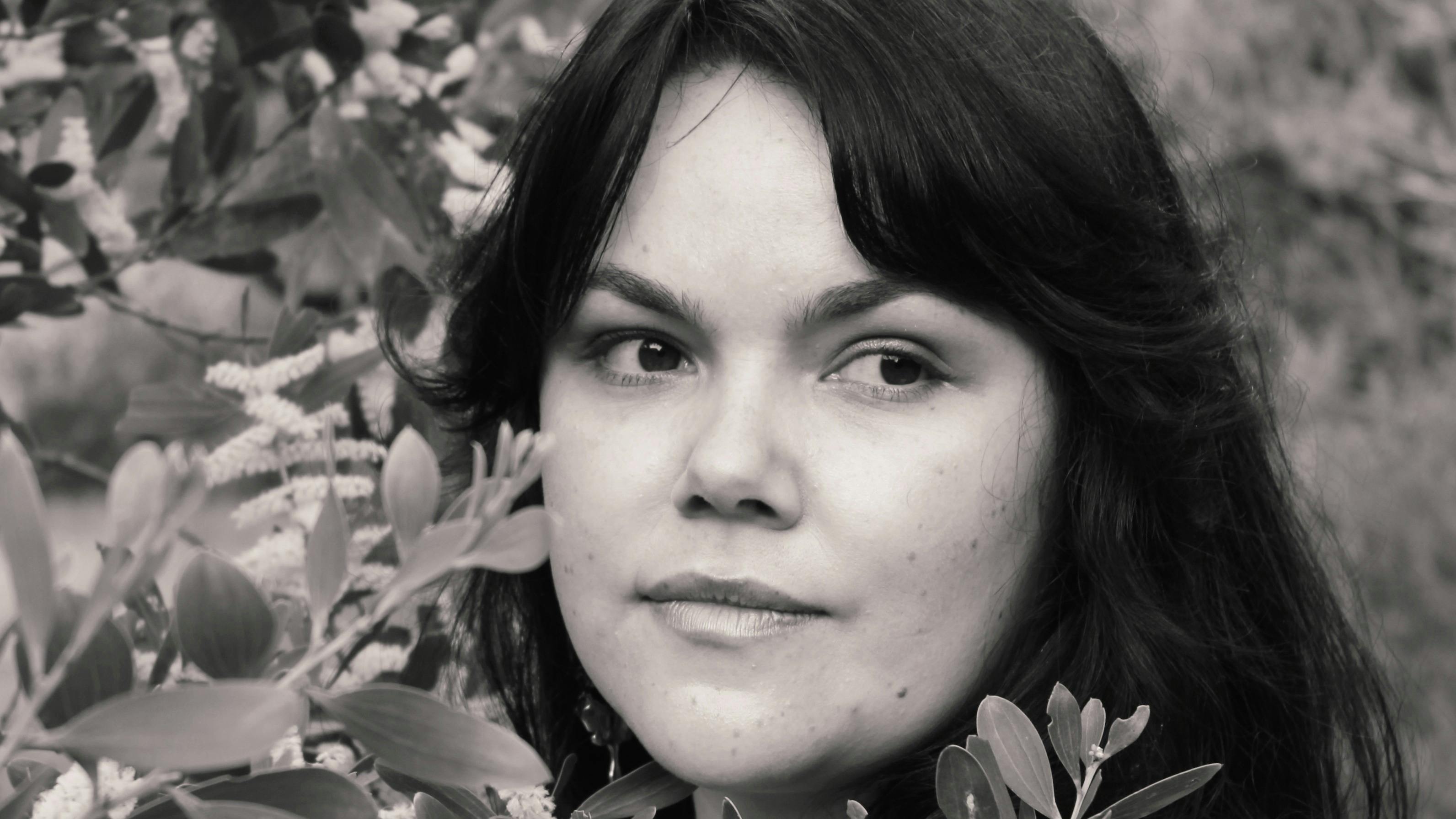A black and white photo of a woman posing beside greenery